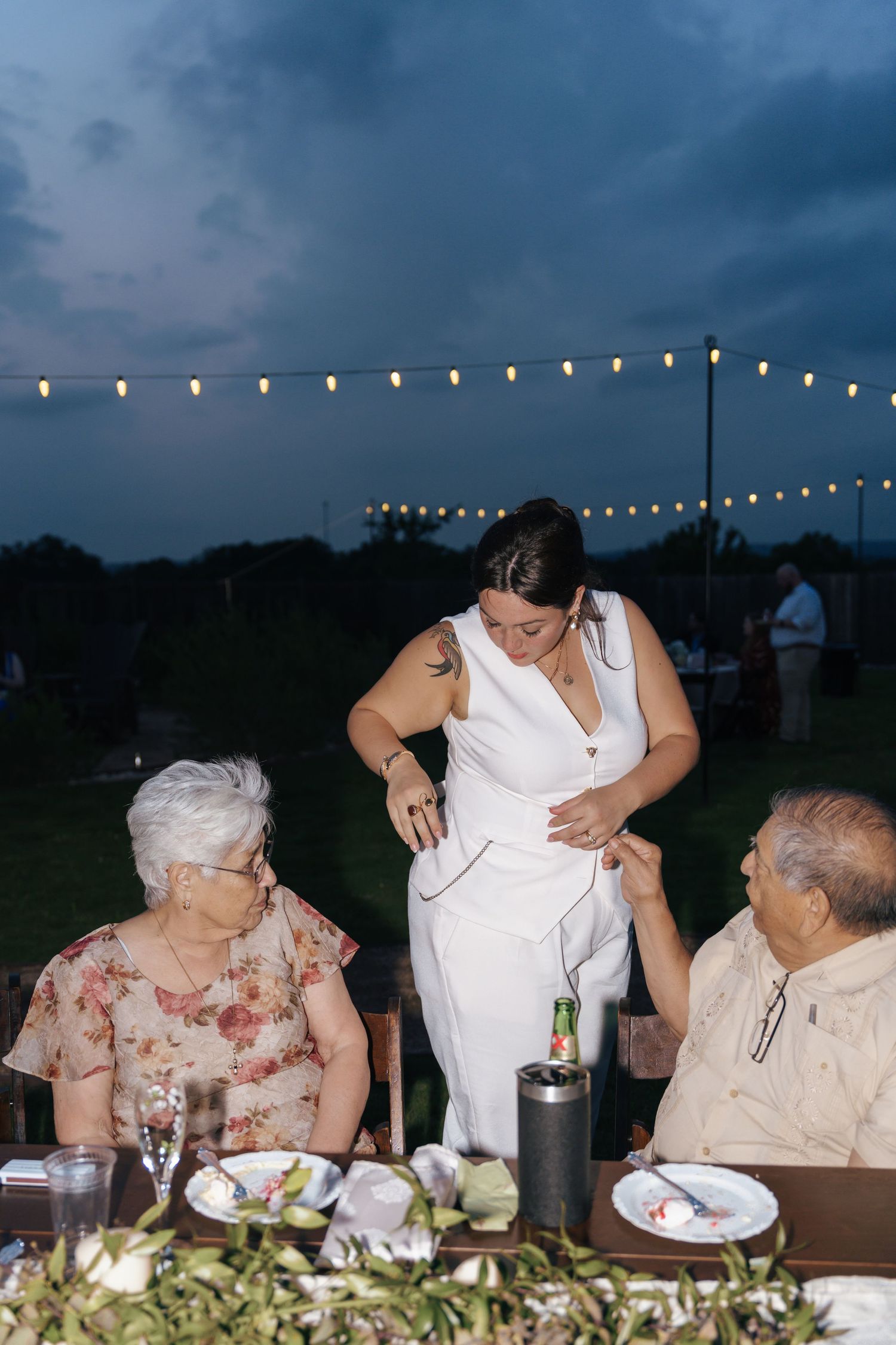 String lights illuminate an outdoor evening gathering as people dine at a long table under darkening skies.