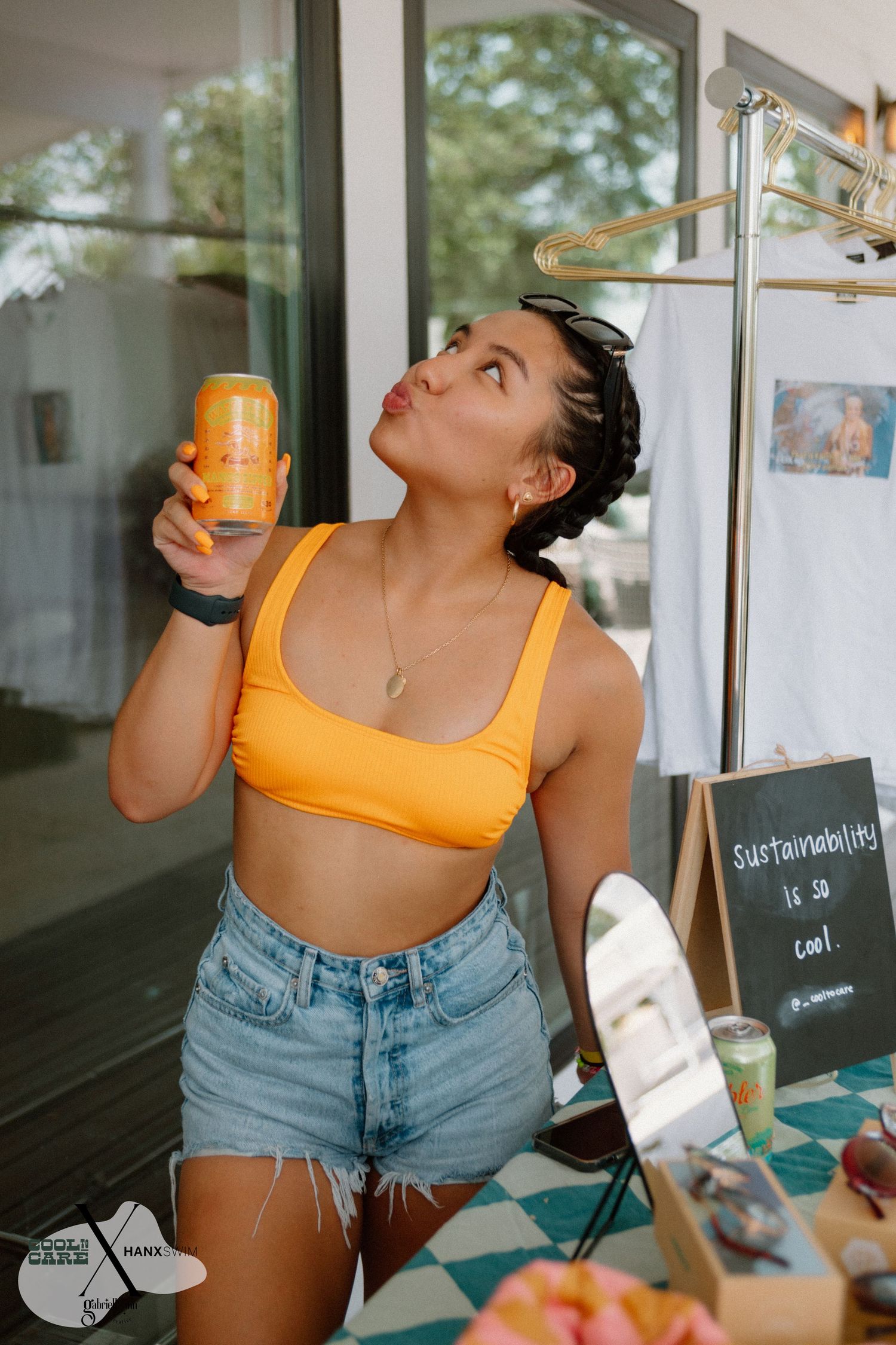 Person in orange top and denim shorts drinking from a beverage container.