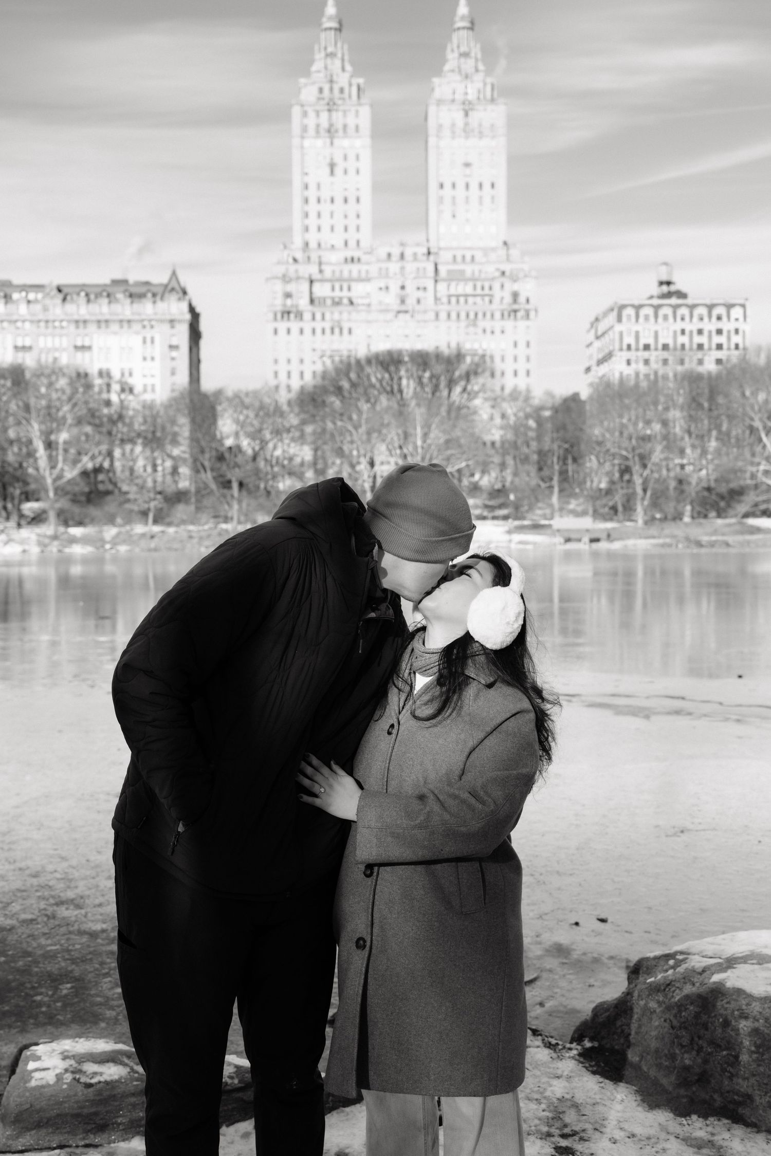 A black and white photograph of two figures by a lake with city buildings visible across the water.