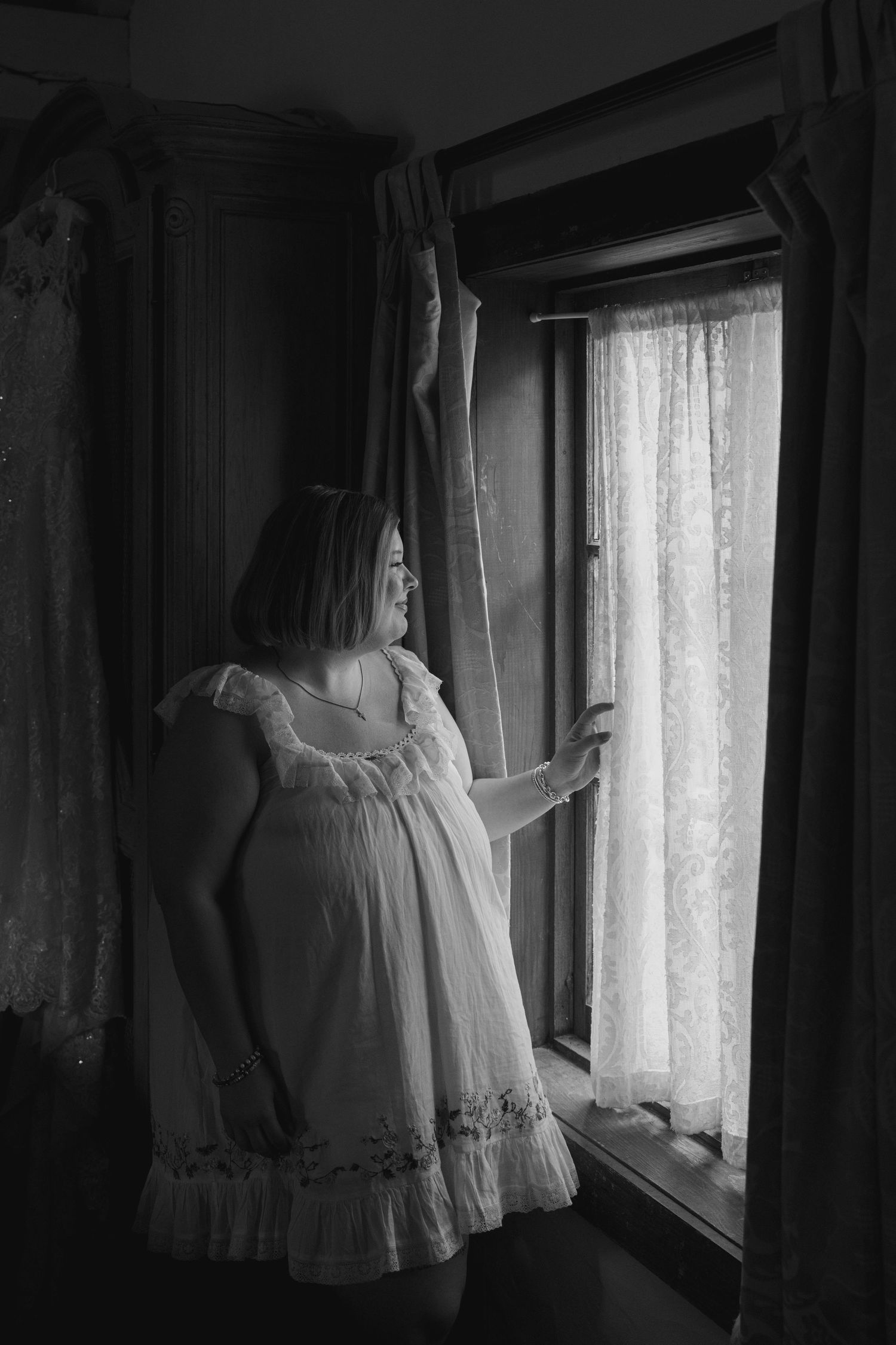 A series of white vintage dresses hanging near a window with lace curtains in moody black and white photography.