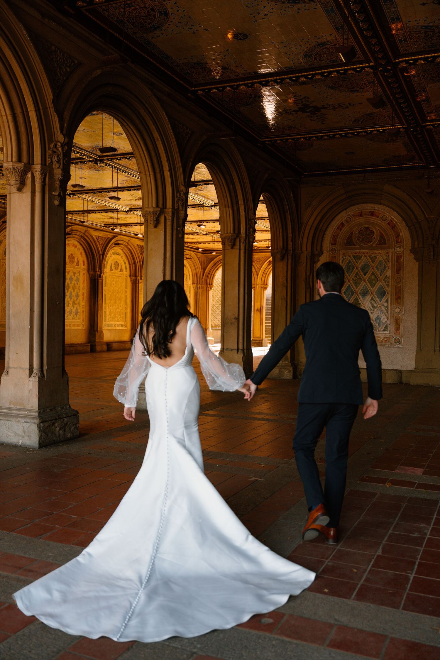 A couple in wedding attire walks through an ornate archway tunnel with beautiful architectural details and warm lighting.