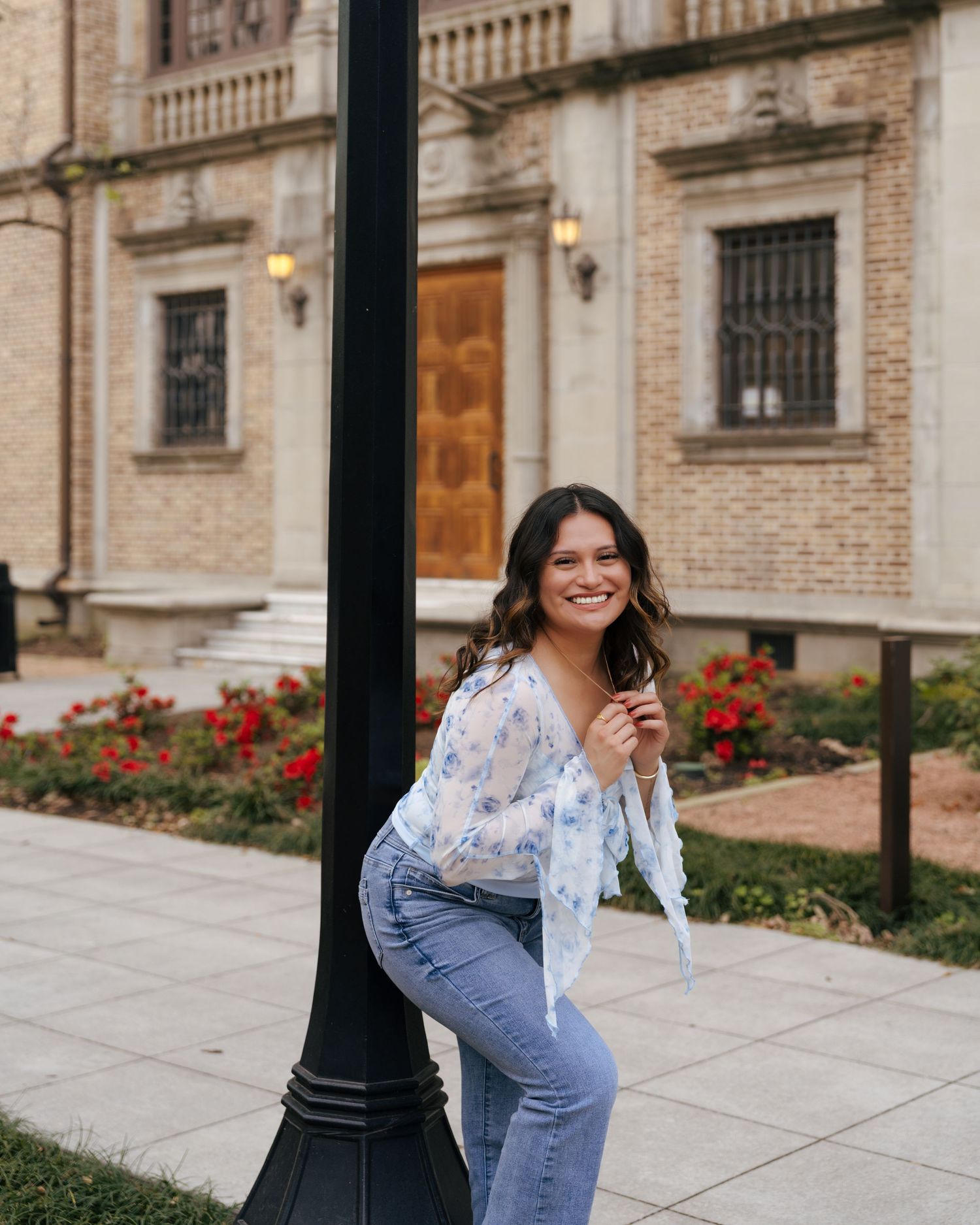 A person in a white floral top and jeans poses cheerfully against a historic stone building with red flowers.