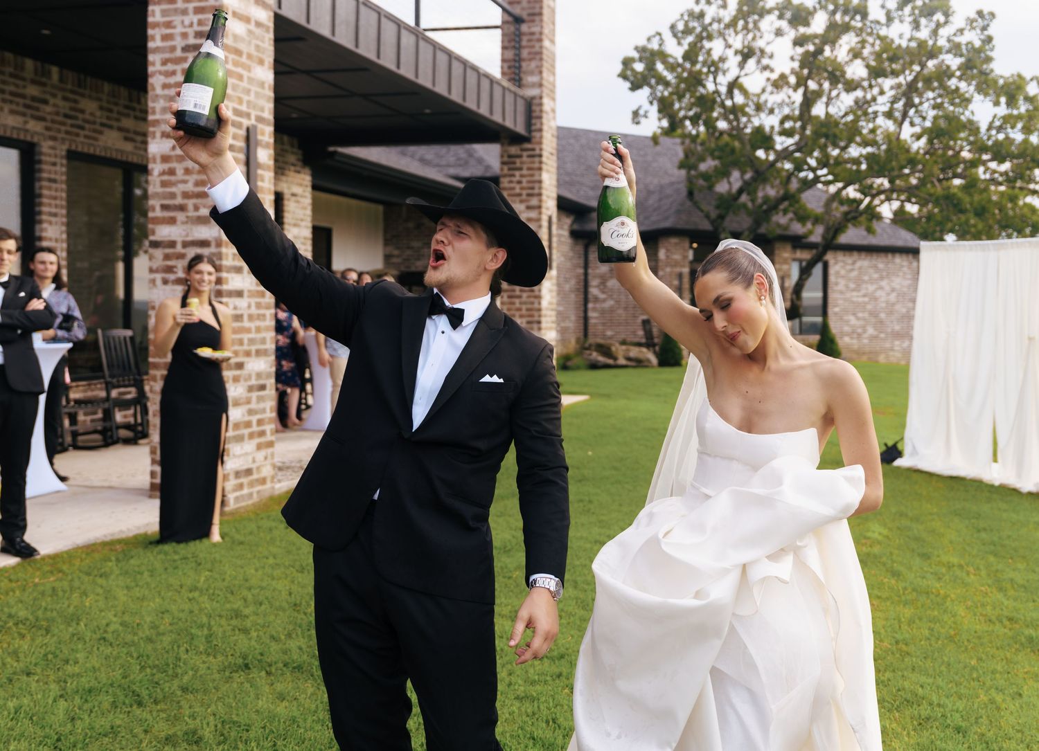 A celebratory toast on the lawn as the newlyweds raise their glasses in front of a brick venue.