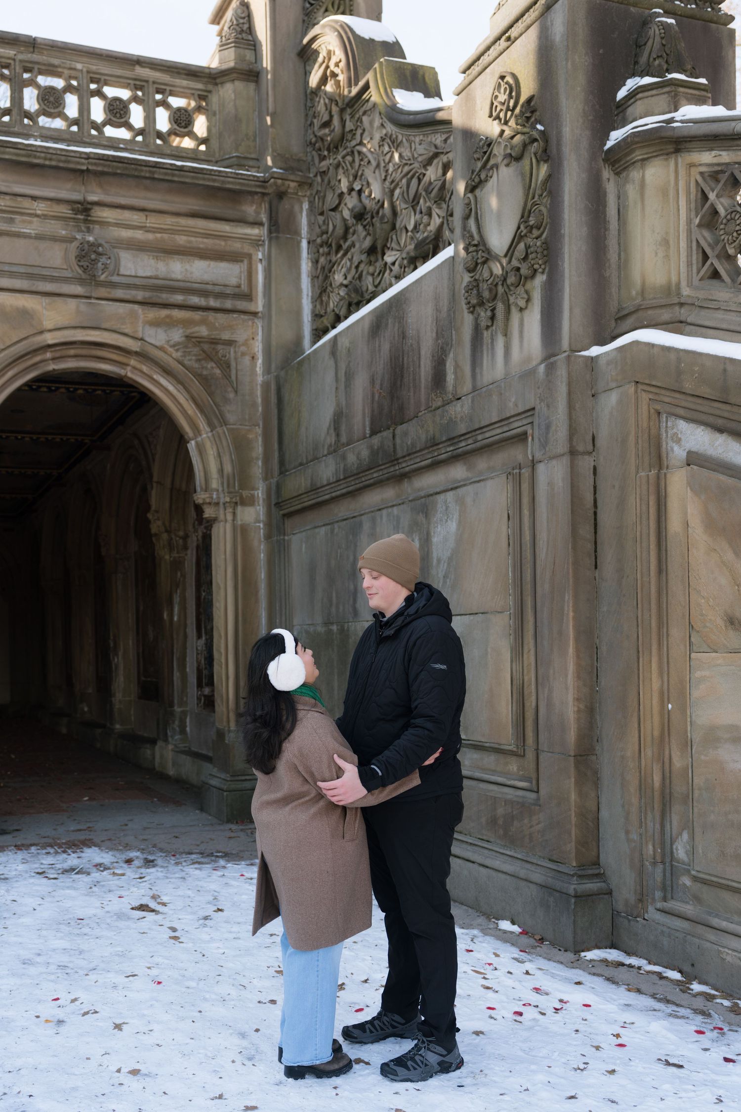A romantic proposal scene in a snowy stone archway with scattered rose petals on the ground.