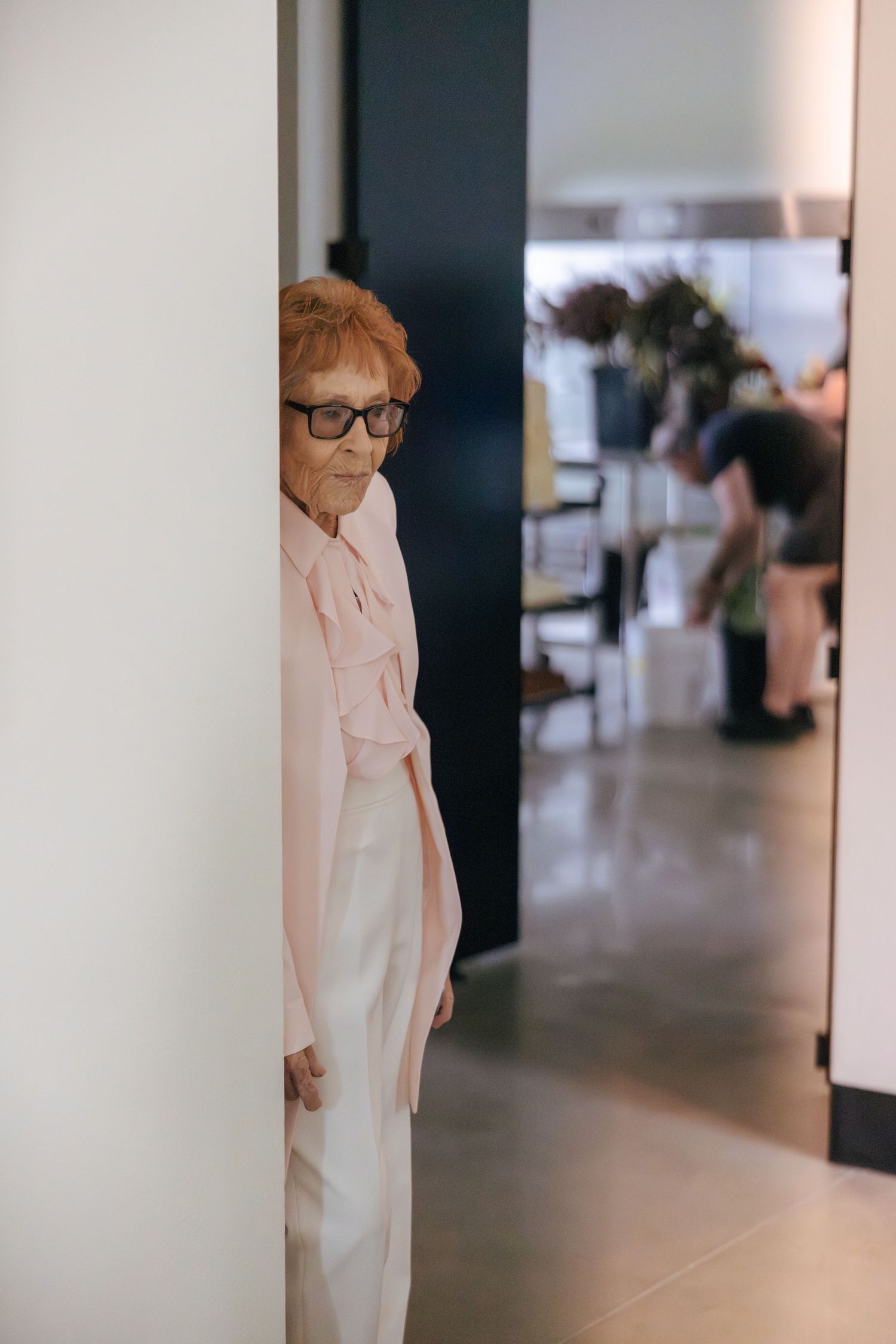 Person in pink blazer and white pants peeking around corner in modern interior hallway.