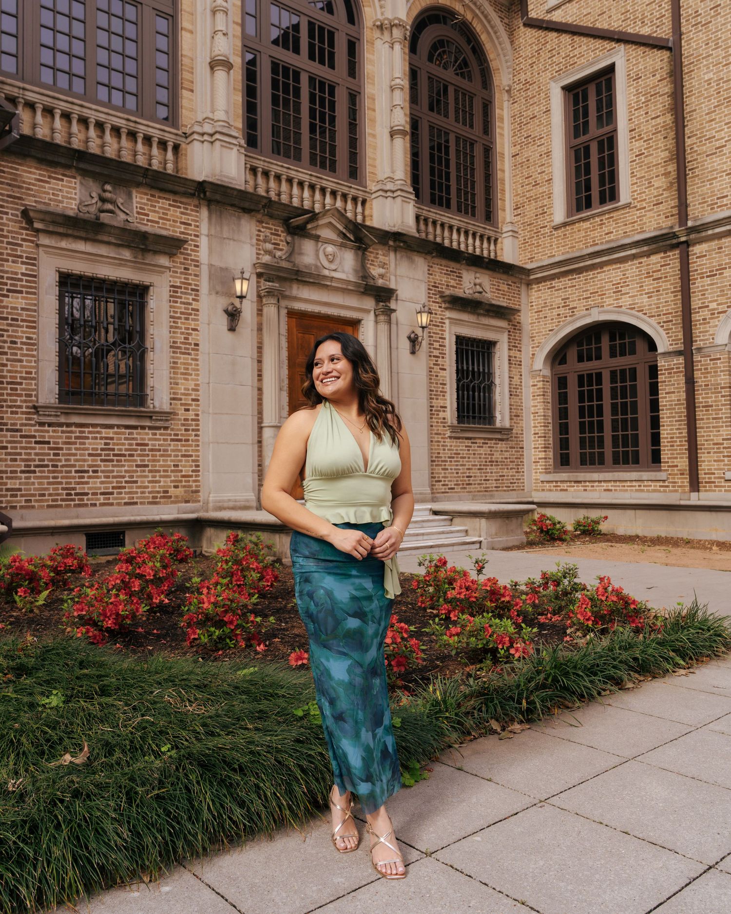 A person in a teal dress poses gracefully in front of a historic brick building with blooming red flowers.