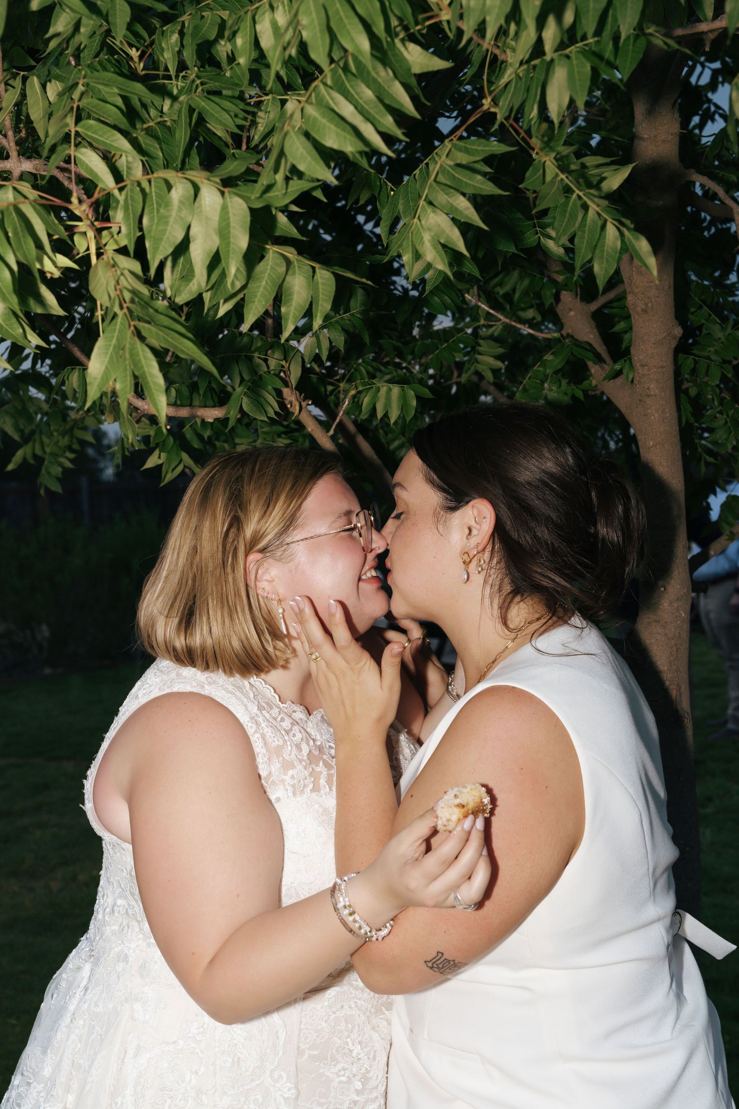 A couple shares a romantic kiss under green leafy trees on their wedding day.