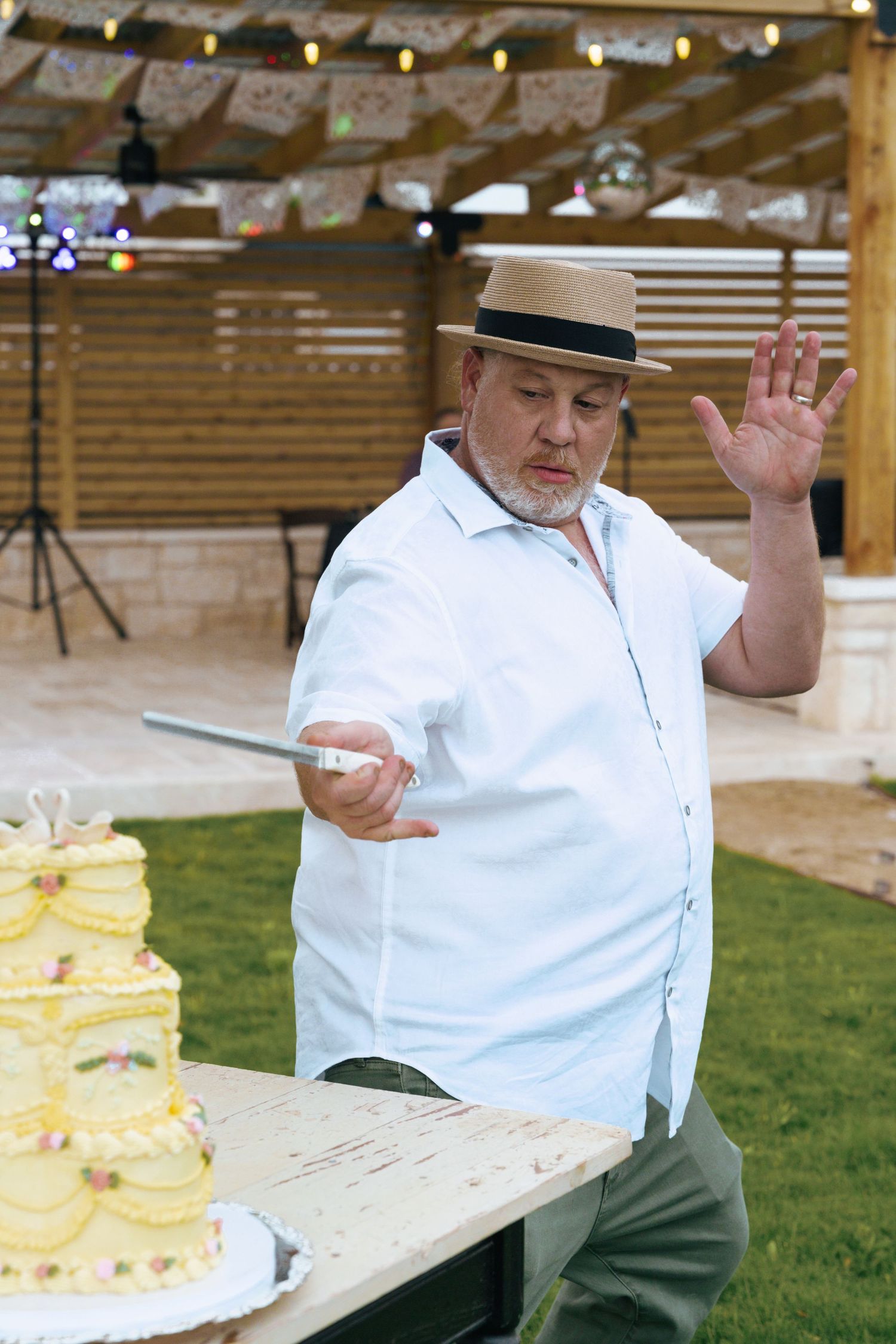 Person in white shirt and hat gestures while sitting at outdoor table with yellow cake slices.
