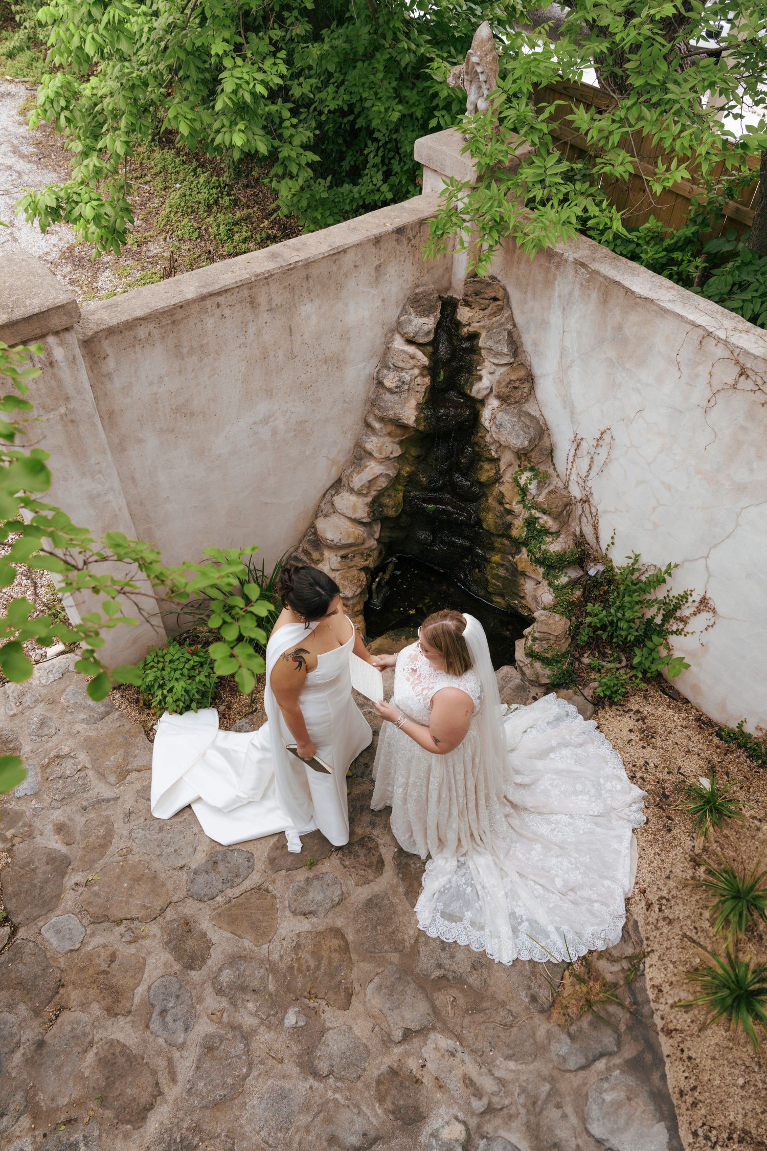 Two wedding dresses elegantly displayed in a rustic stone courtyard with green foliage overhead.