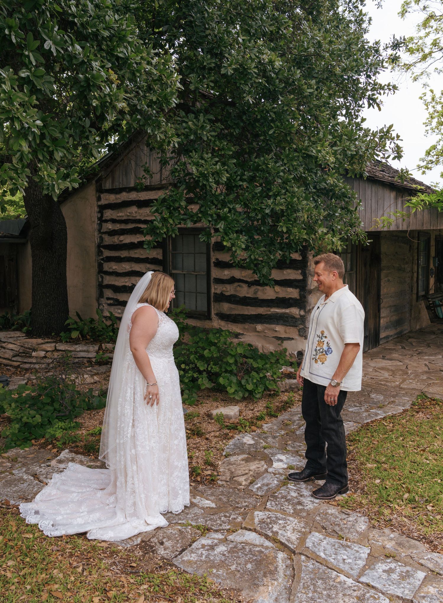 A couple stands in front of a rustic log cabin with a woman wearing a flowing white lace dress on a stone path.