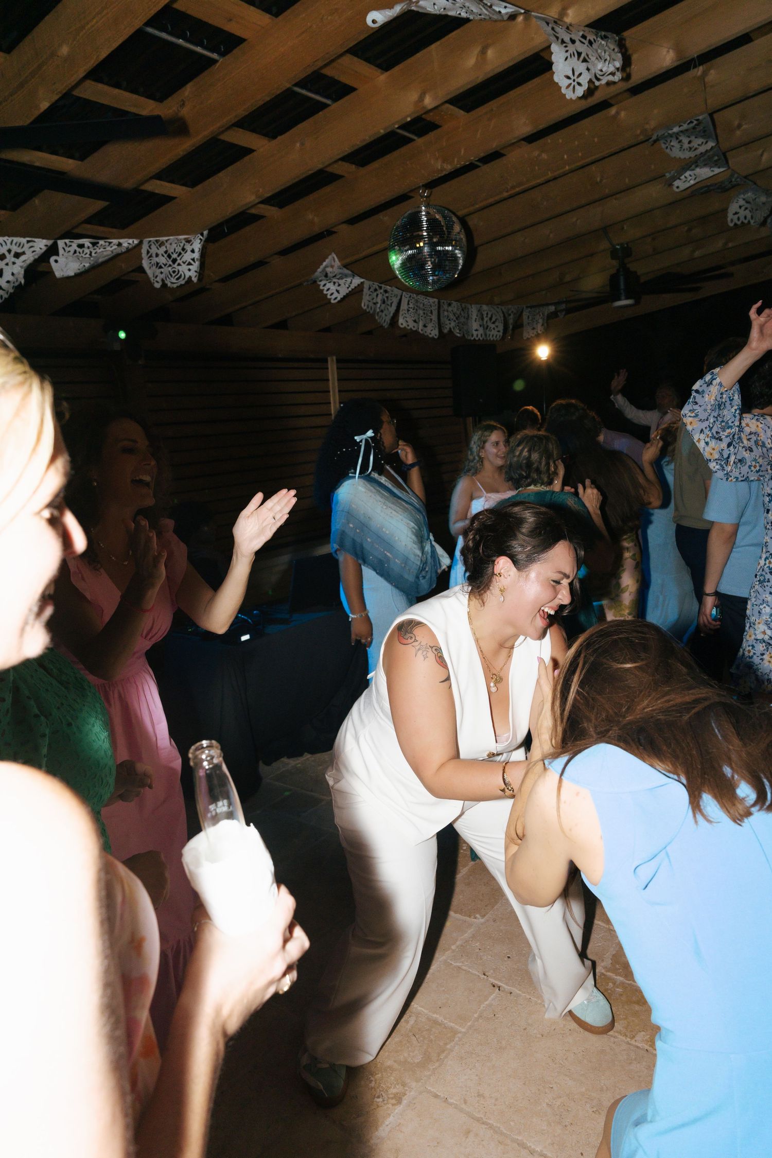 People dancing and celebrating at a party under wooden beams decorated with white banners and lights.