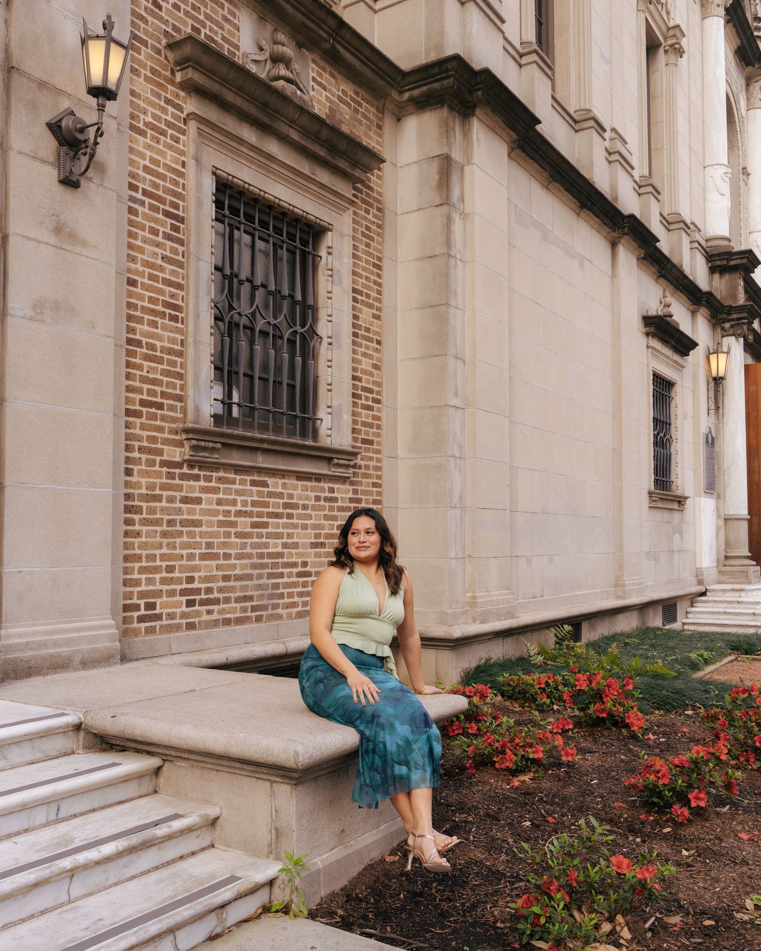 Person in a teal dress poses elegantly on stone steps next to red flowers and brick walls of a historic building.