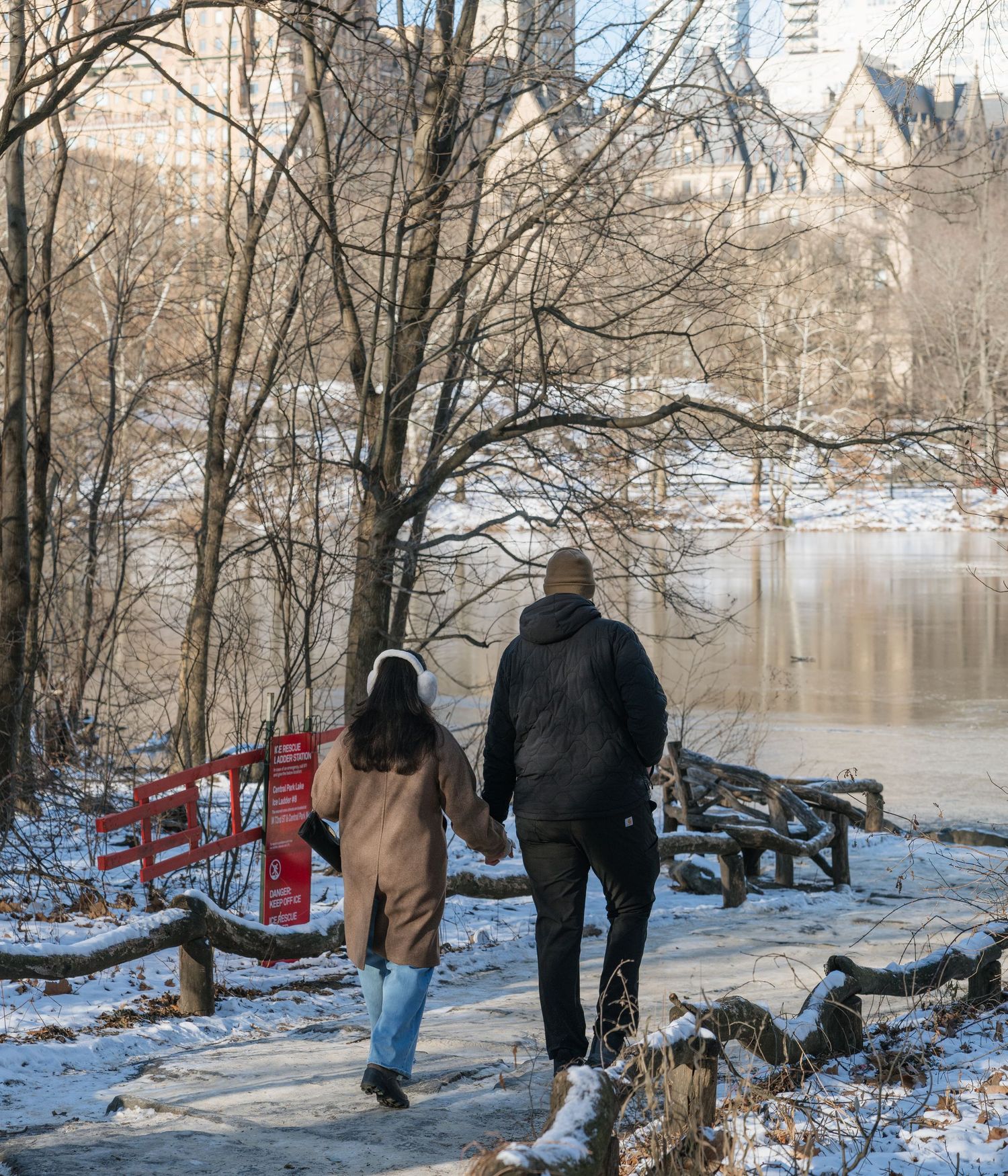 People walking along a snowy riverbank on a winter day with bare trees and water in the background.