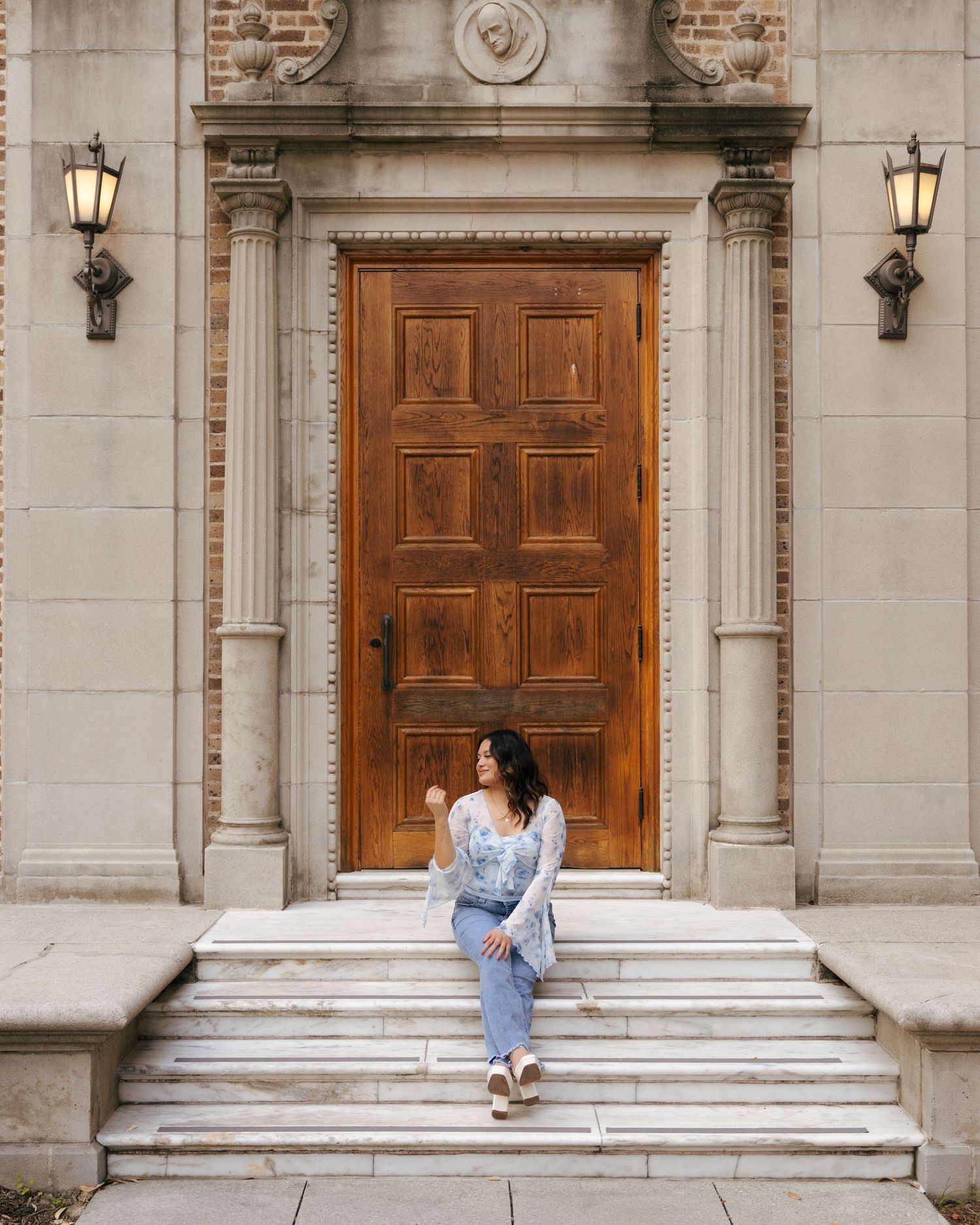 A large wooden door with ornate stone columns and lanterns frames someone sitting on stone steps in casual attire.