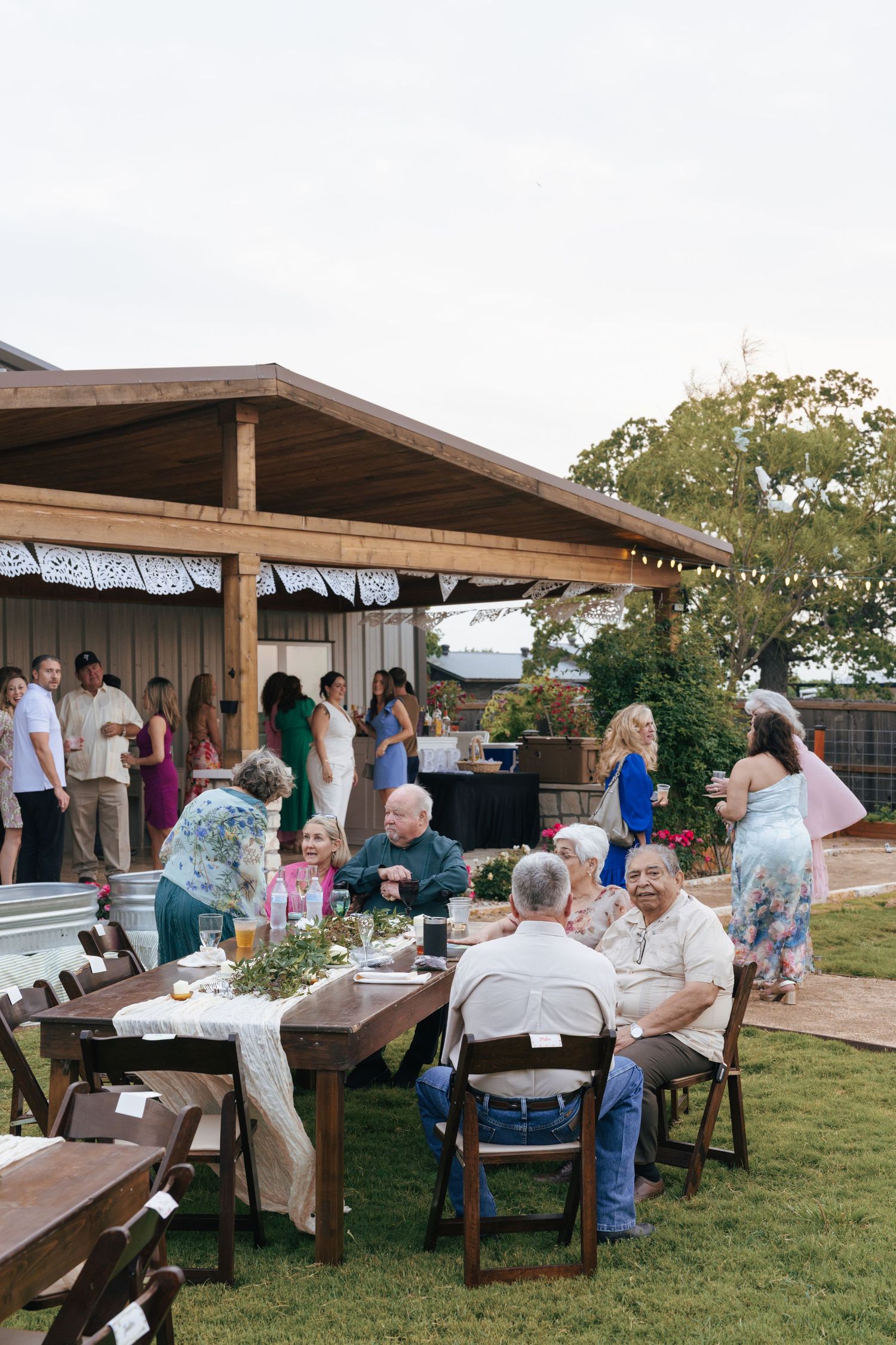 People gather at outdoor picnic tables under wooden pavilions for a casual community event.