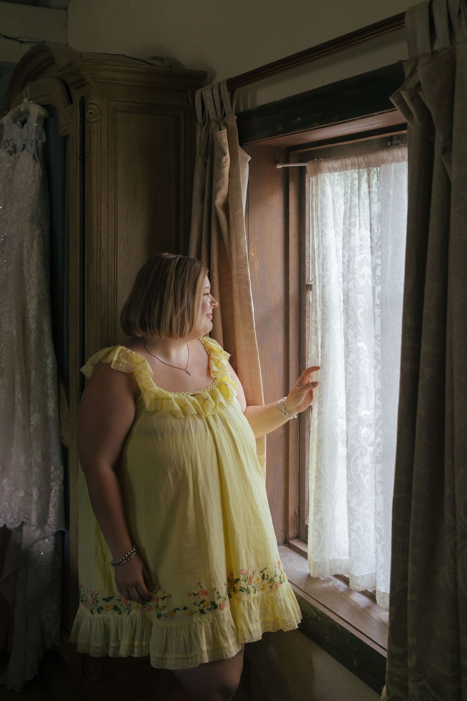 A figure in a yellow vintage-style dress stands by a window with lace curtains in a dimly lit room.