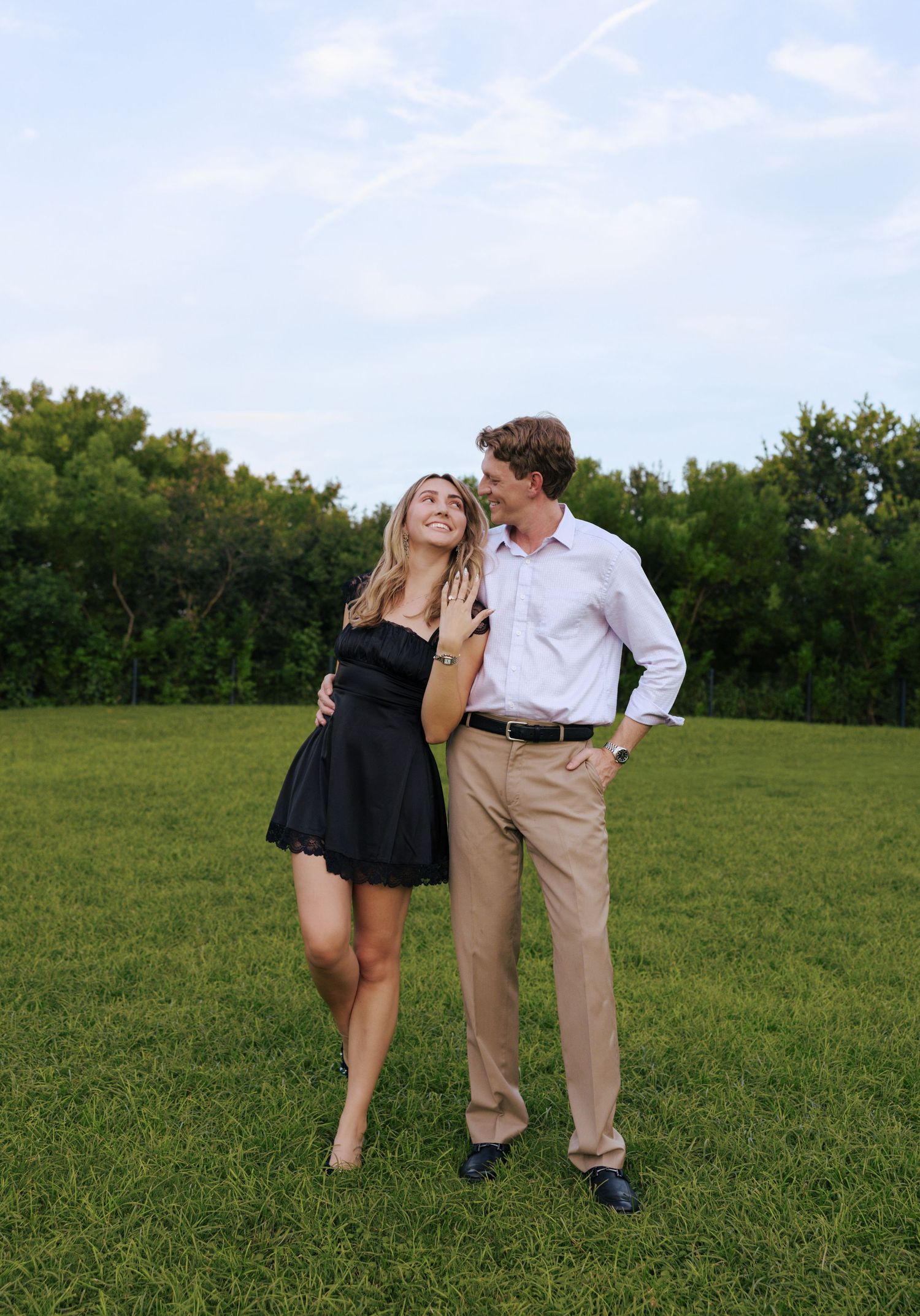 A couple in formal attire shares a romantic moment together on a grassy field with trees in the background.
