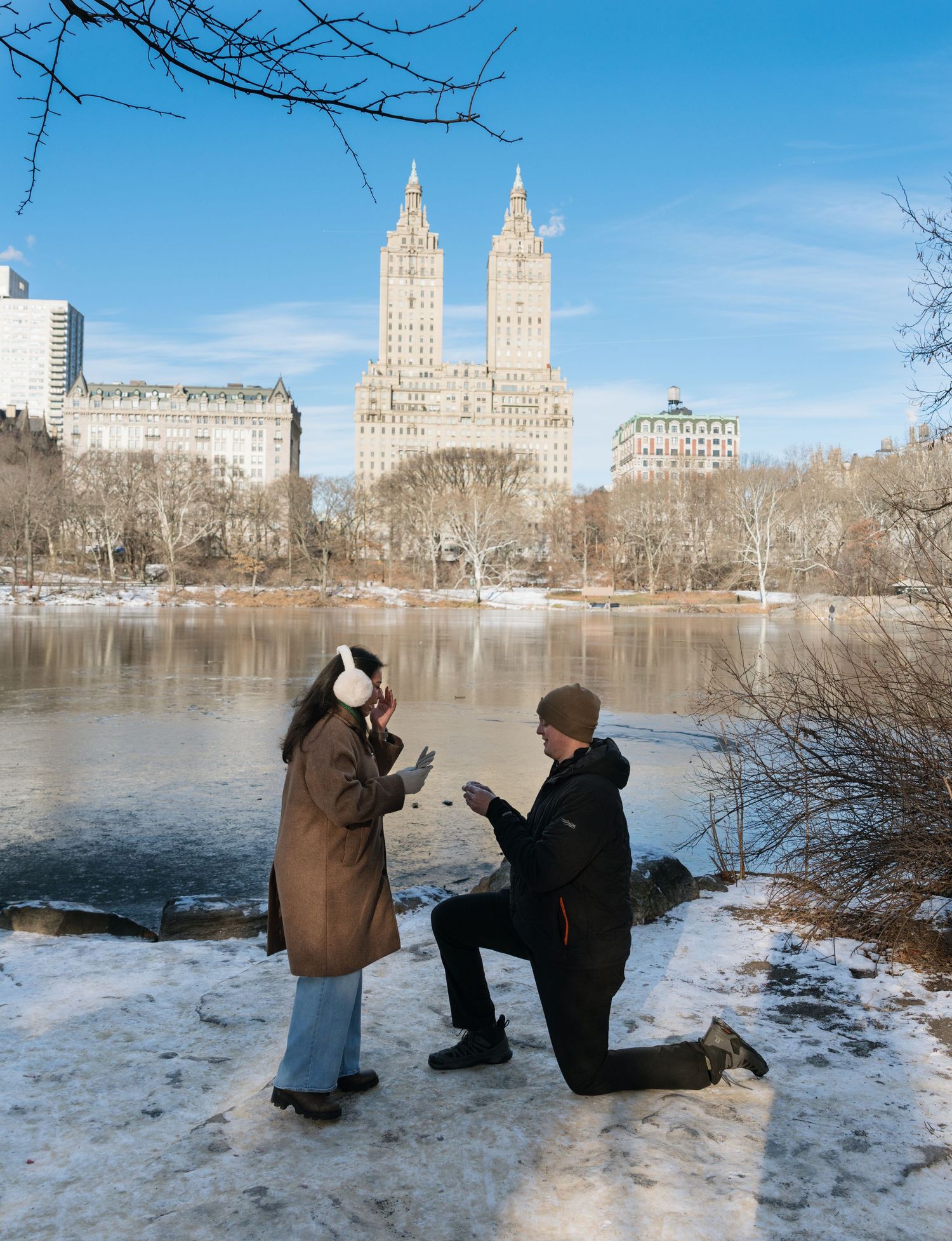 People photographing a winter scene at Central Park Lake with iconic New York City buildings in the background.