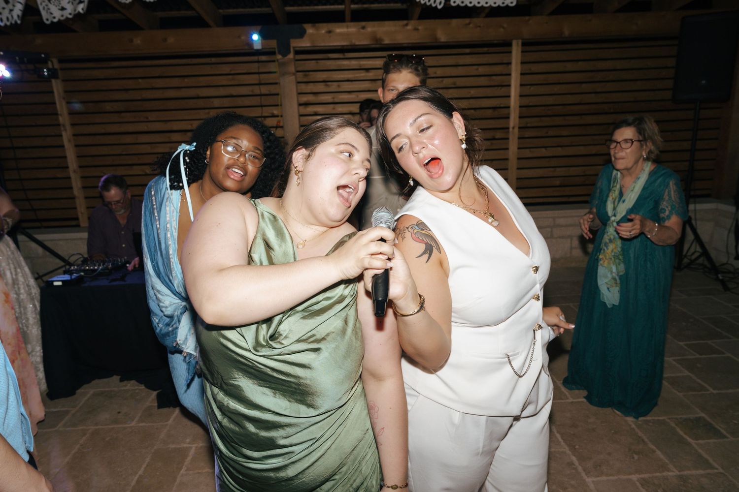 People dance and celebrate at a lively party with wooden walls and dim lighting in the background.