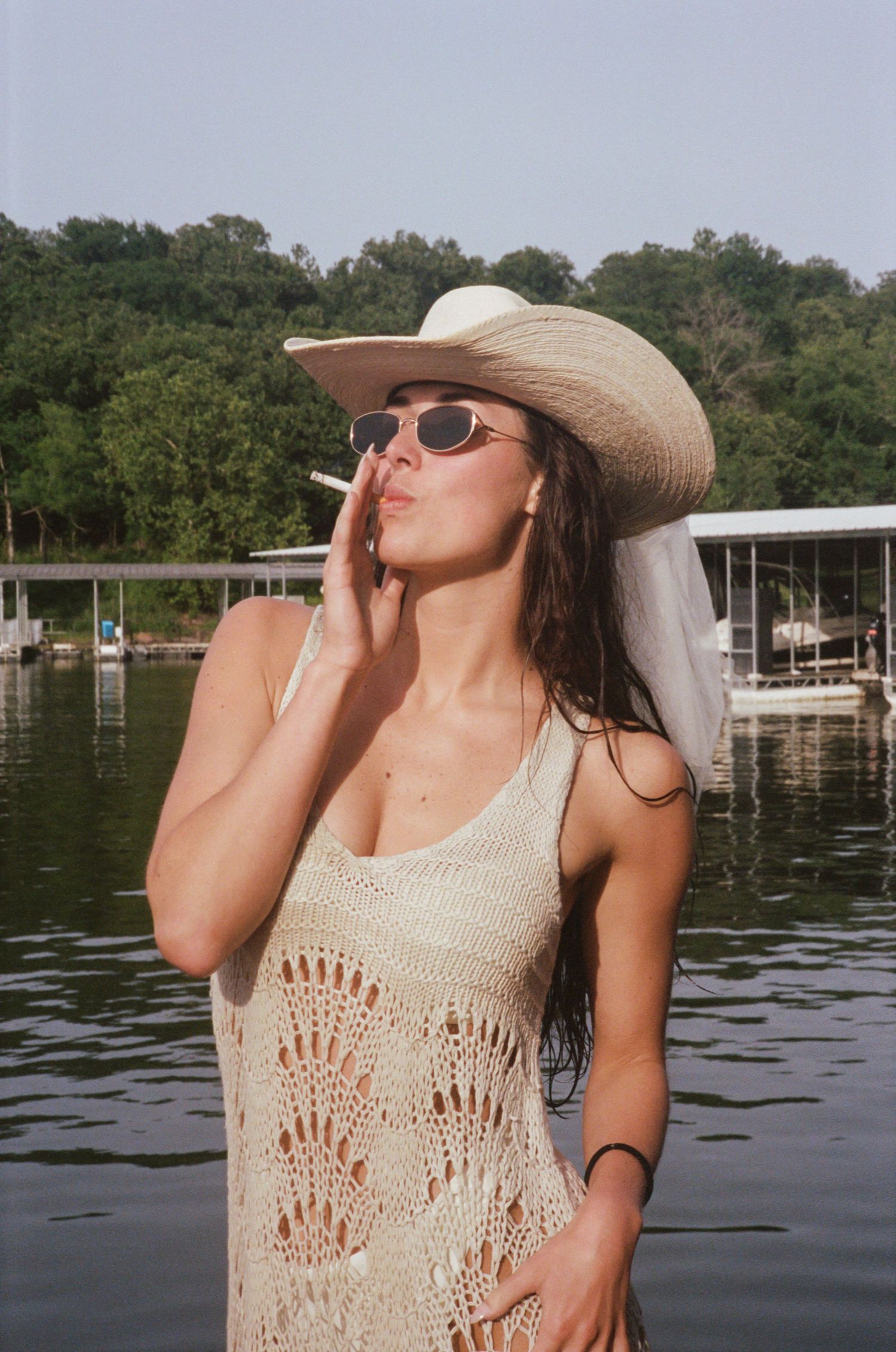 Person in a cream crochet dress and sun hat poses on a boat near a lake with trees in the background.