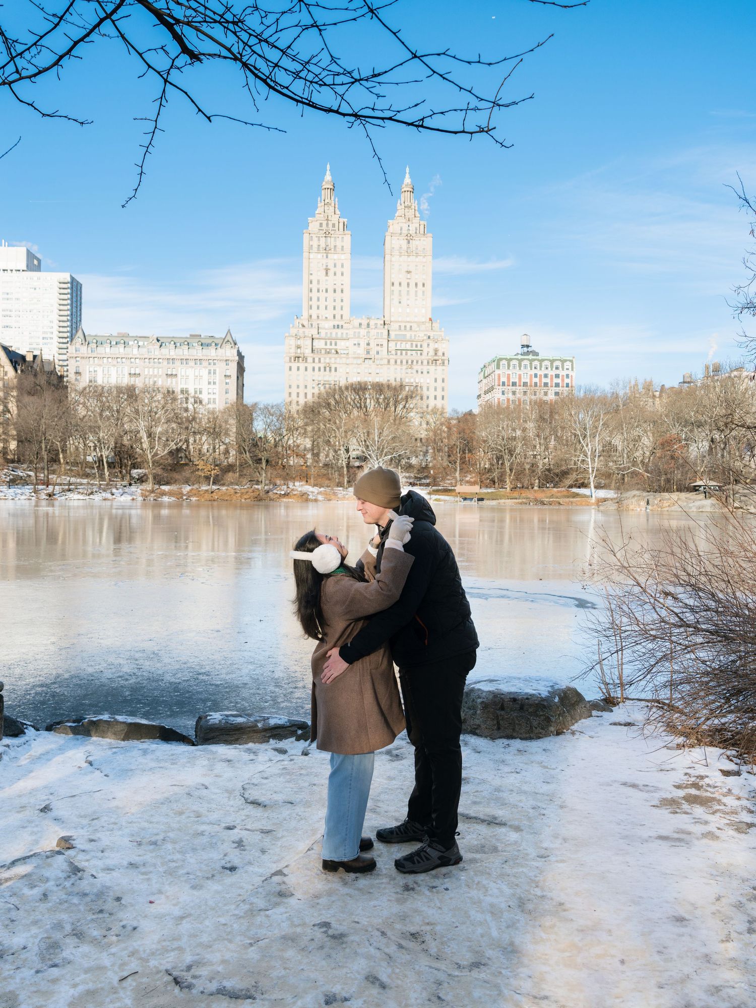 Snowy winter scene in Central Park with iconic New York City buildings visible across the lake.