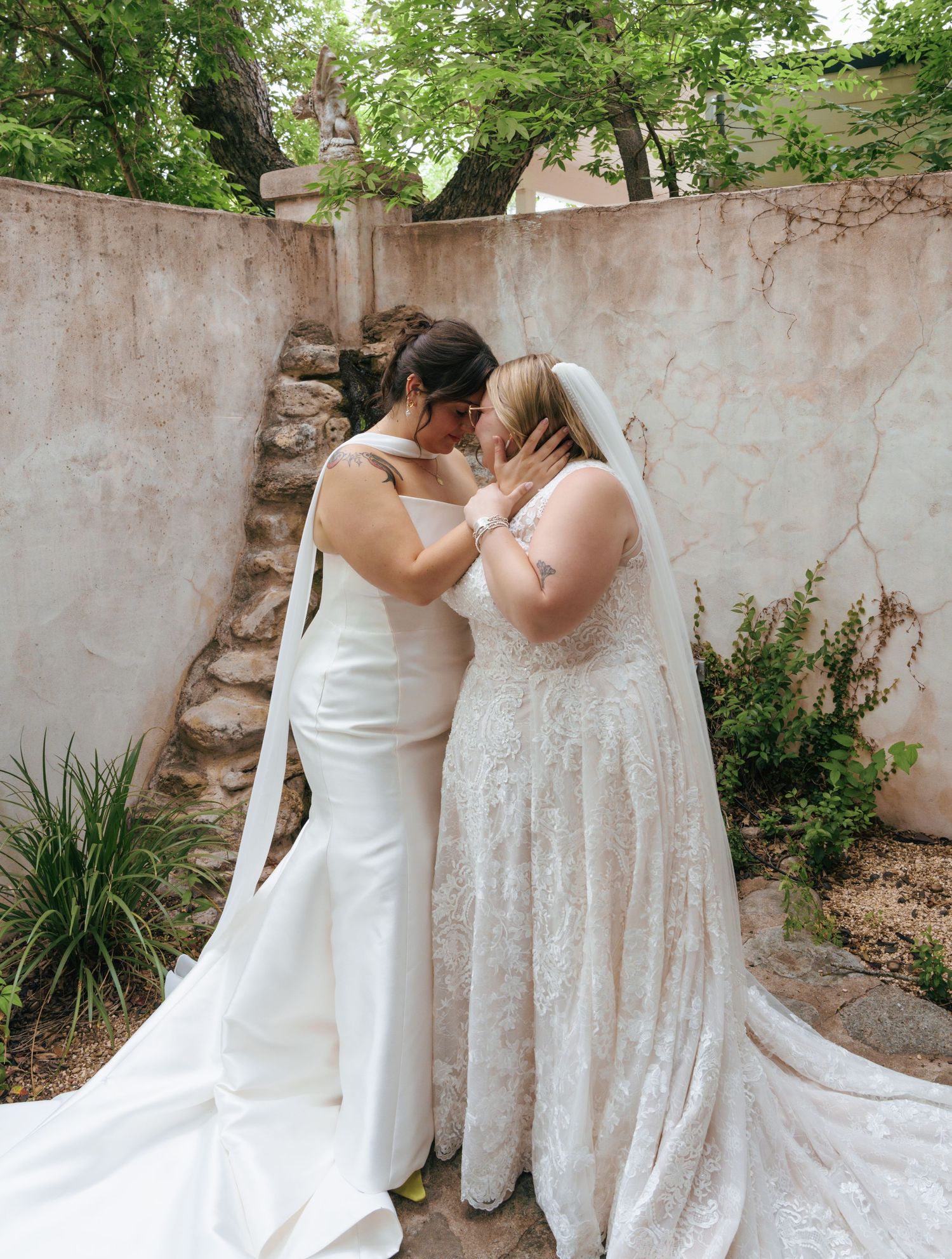 Two people in white wedding dresses pose together against a rustic wall in an outdoor garden setting.