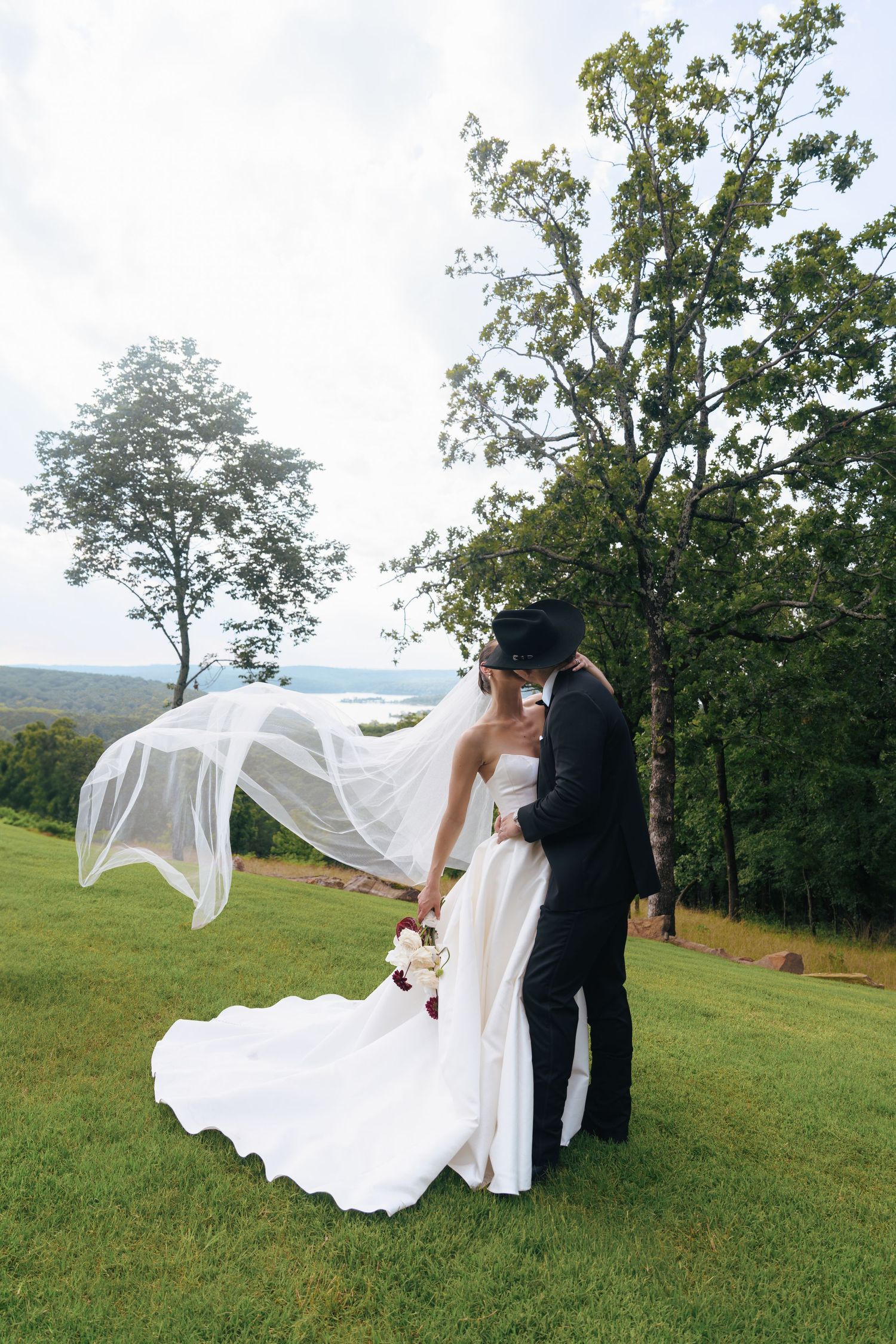 A wedding couple poses on a hilltop as the bride's veil dramatically flows in the wind against a scenic landscape.