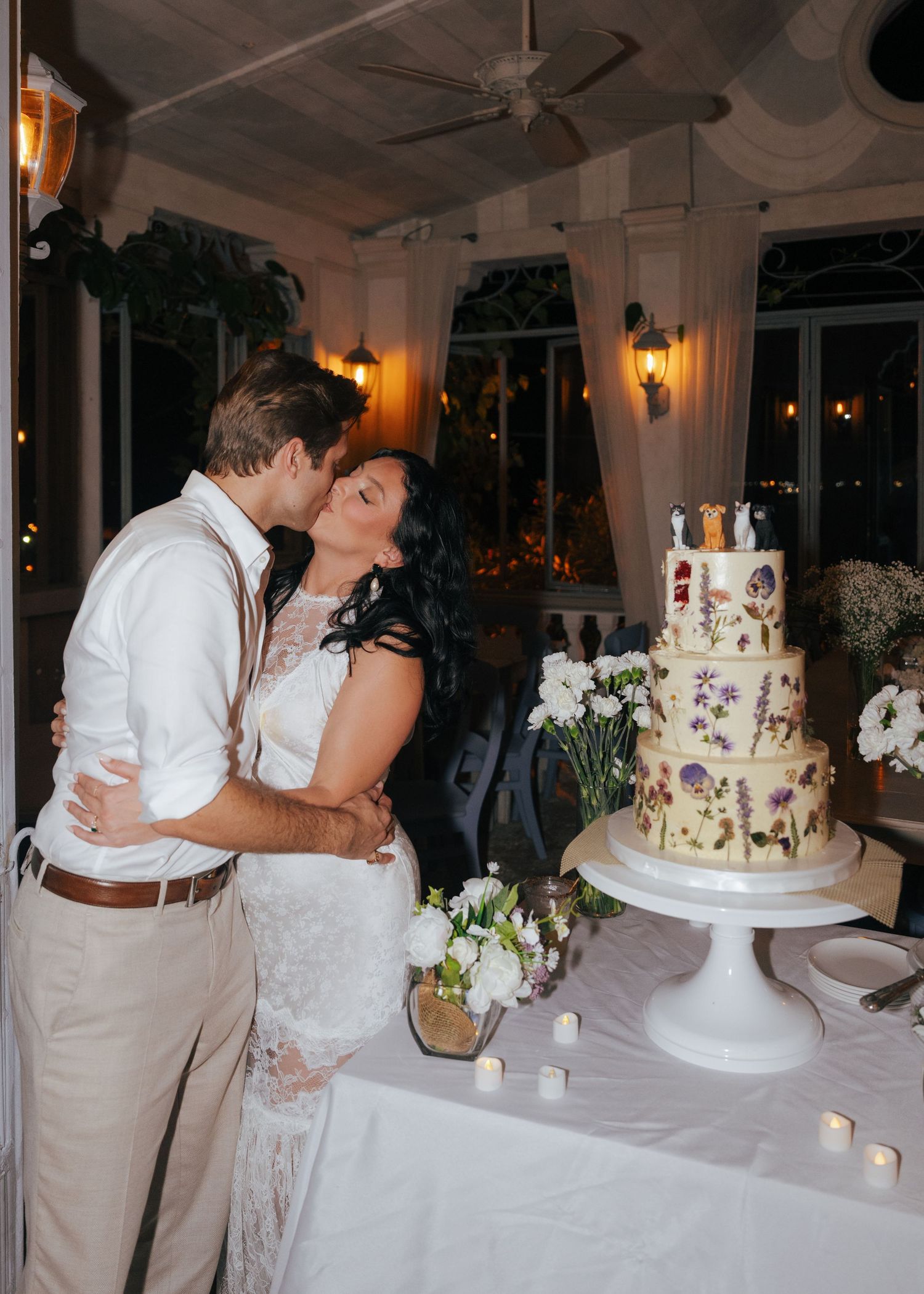 Couple shares romantic kiss by elegant floral wedding cake at evening reception.