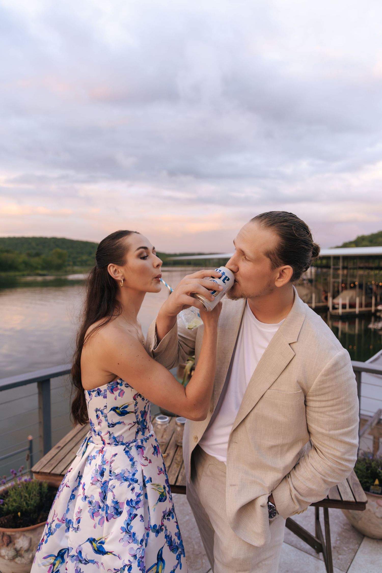 A couple shares an intimate moment on a waterfront deck at sunset while enjoying drinks together.