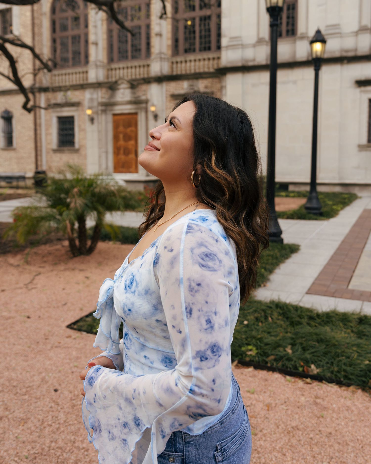 Woman in floral top and overalls laughs joyfully while standing in a garden courtyard with historic architecture.