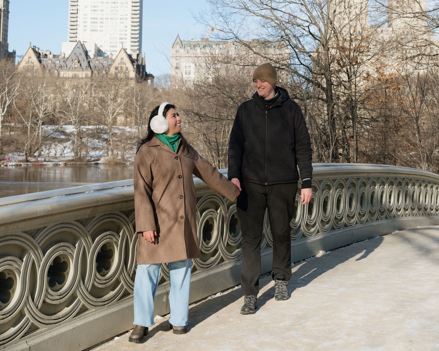 People walking along Bow Bridge in Central Park during winter with snow covered railings and city skyline.