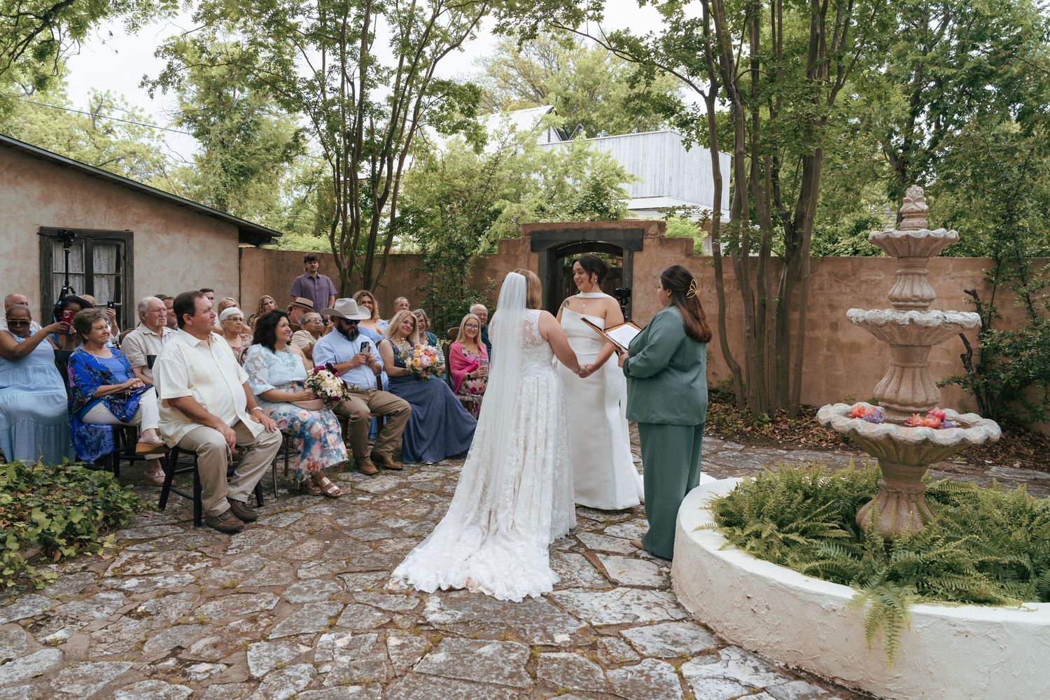 An intimate outdoor wedding ceremony takes place in a courtyard garden with a stone fountain and guests seated on benches.