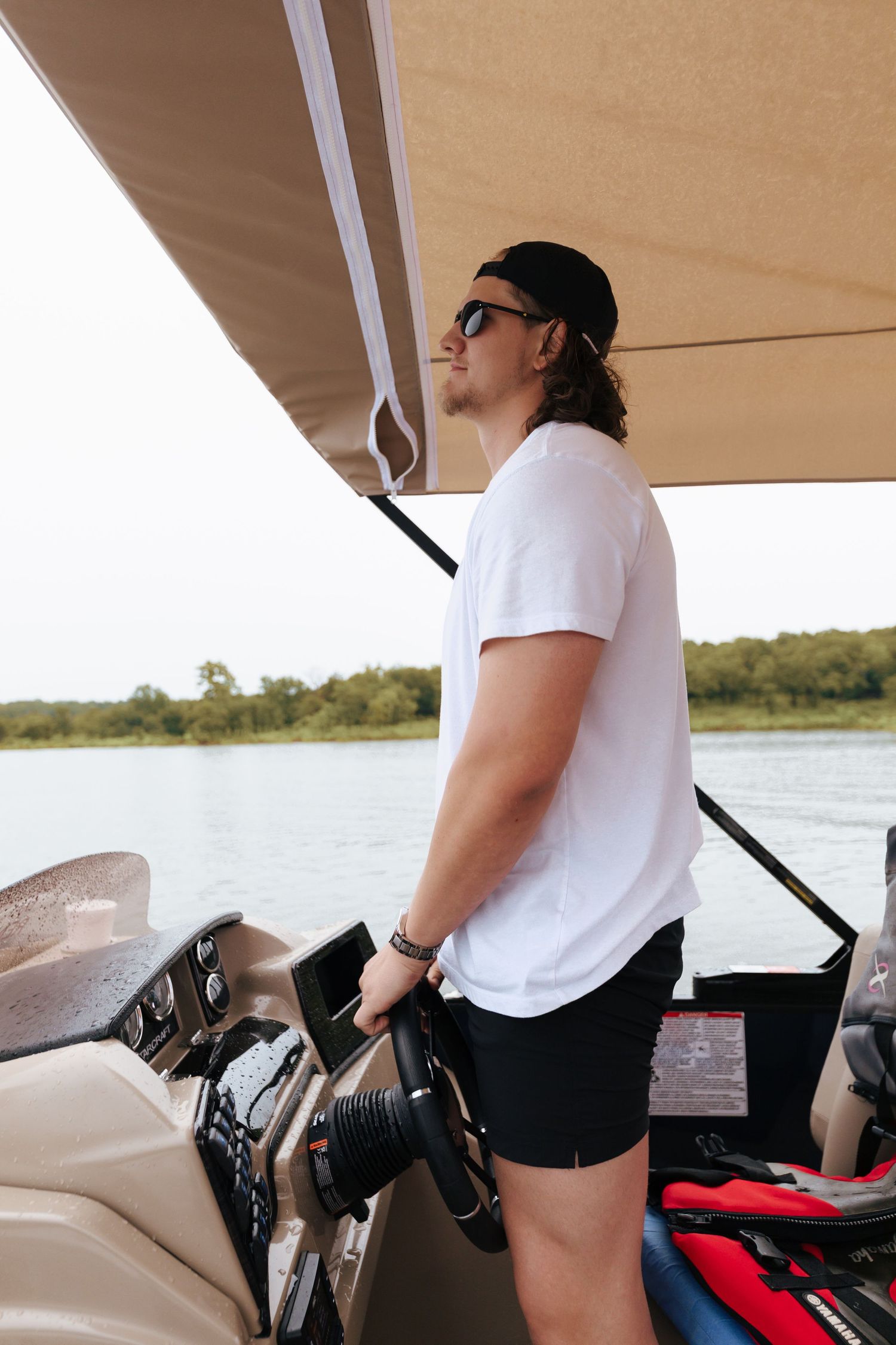 Person in white t-shirt and black shorts steering a boat on calm waters under a sunny sky.