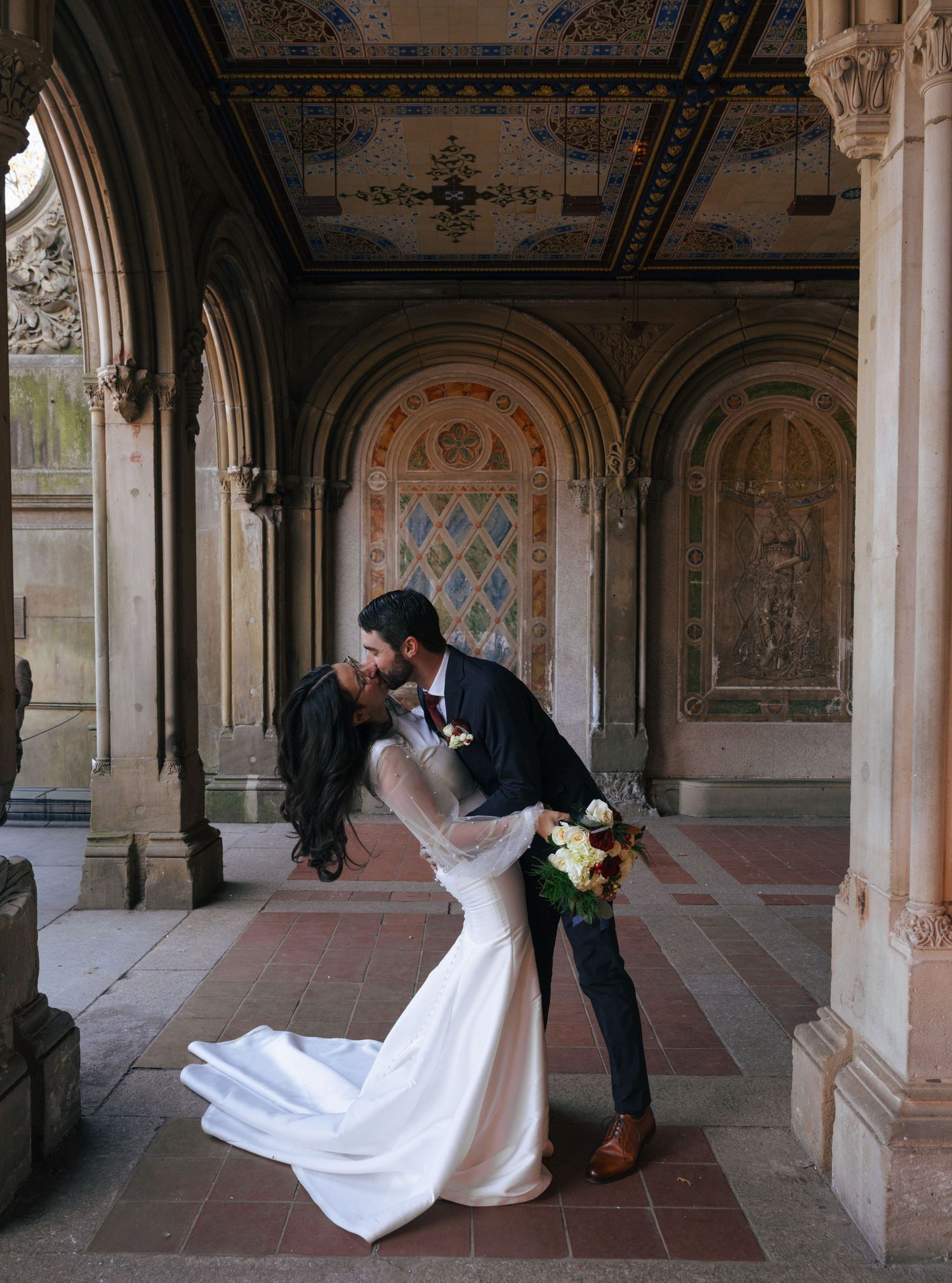 A romantic wedding couple shares a kiss under ornate archways at Bethesda Terrace in Central Park.