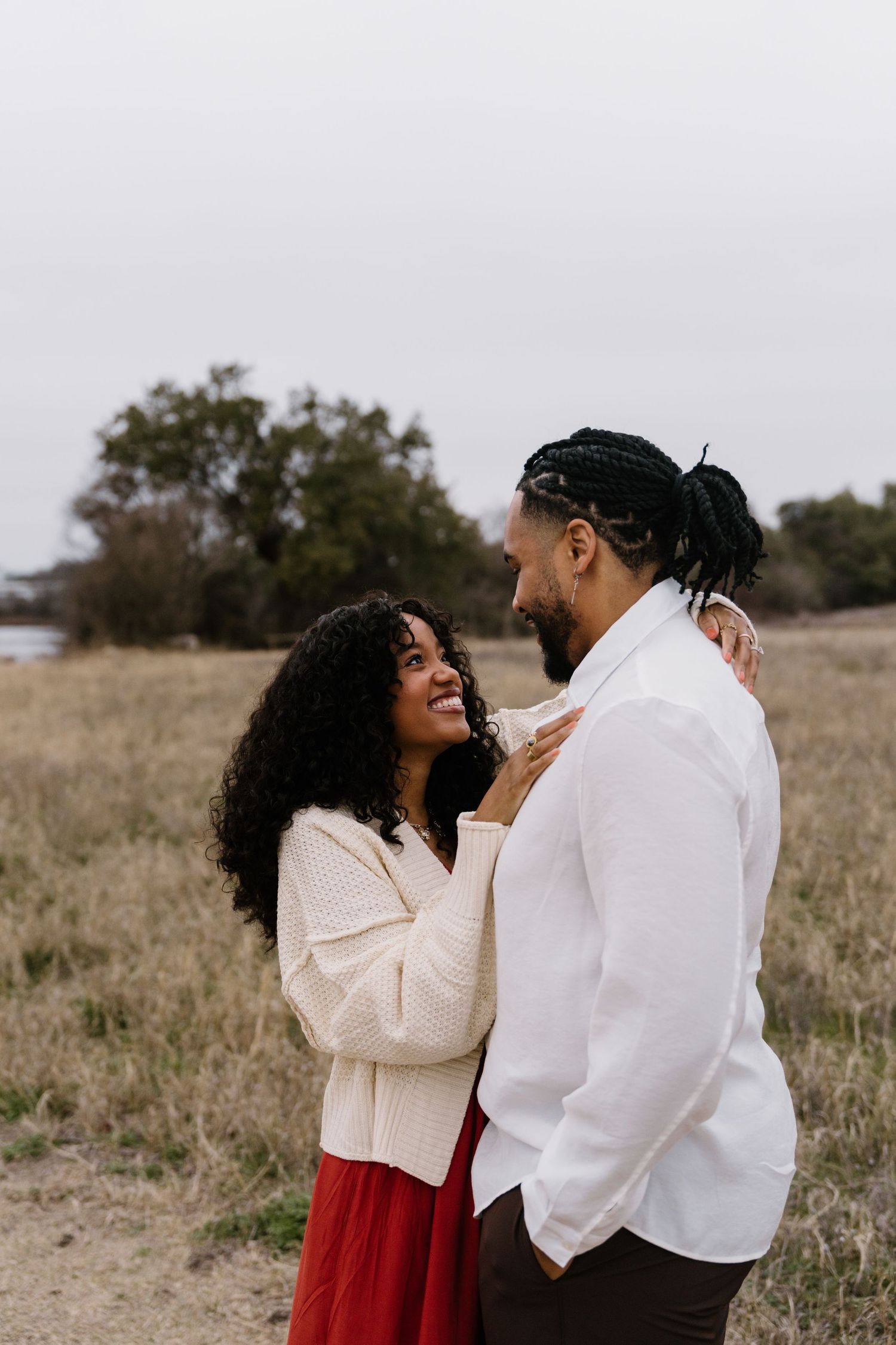 Couple embraces and laughs together in casual attire against rustic field backdrop.
