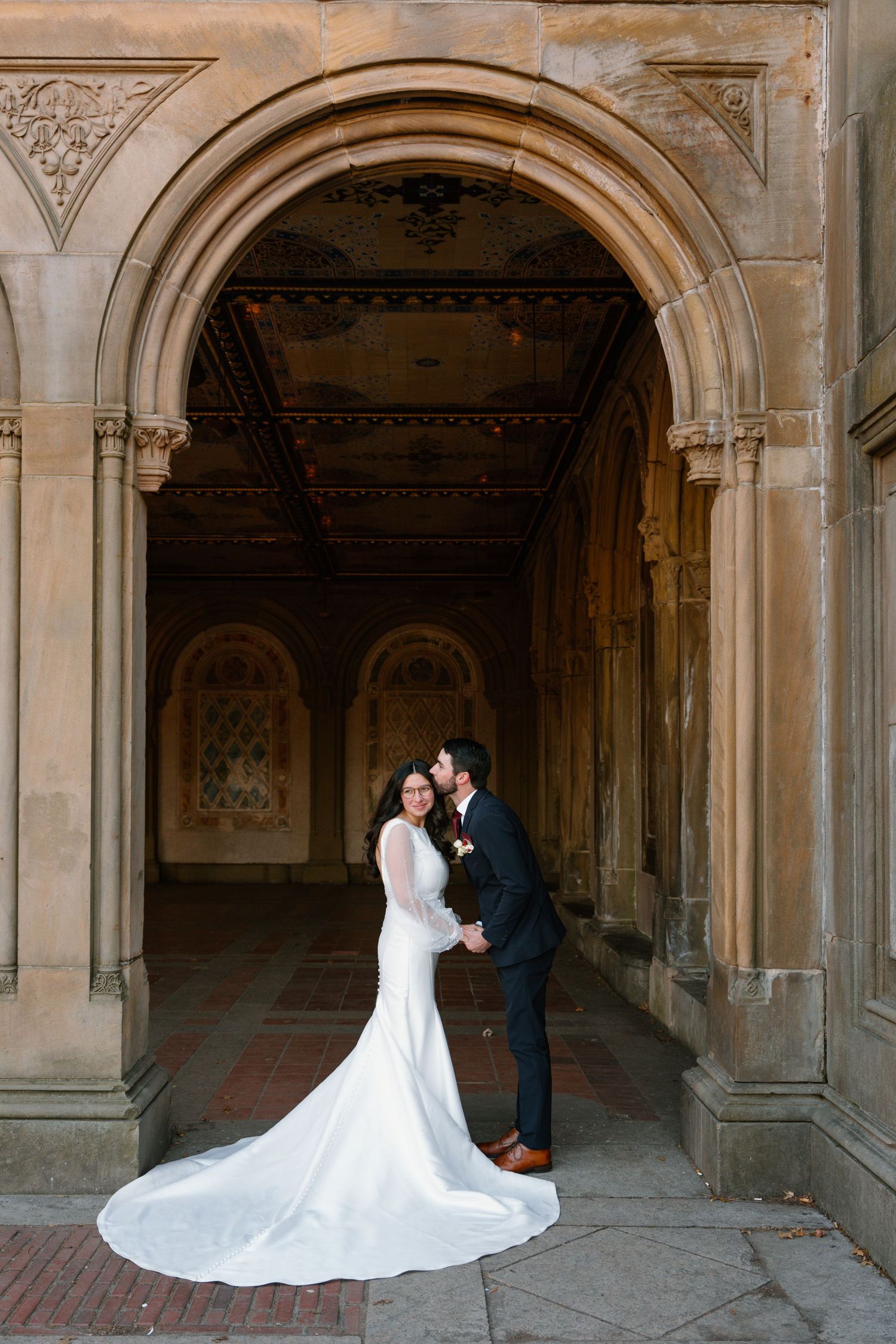 A wedding couple poses beneath ornate stone archways in a historic architectural setting with dramatic lighting and shadows.