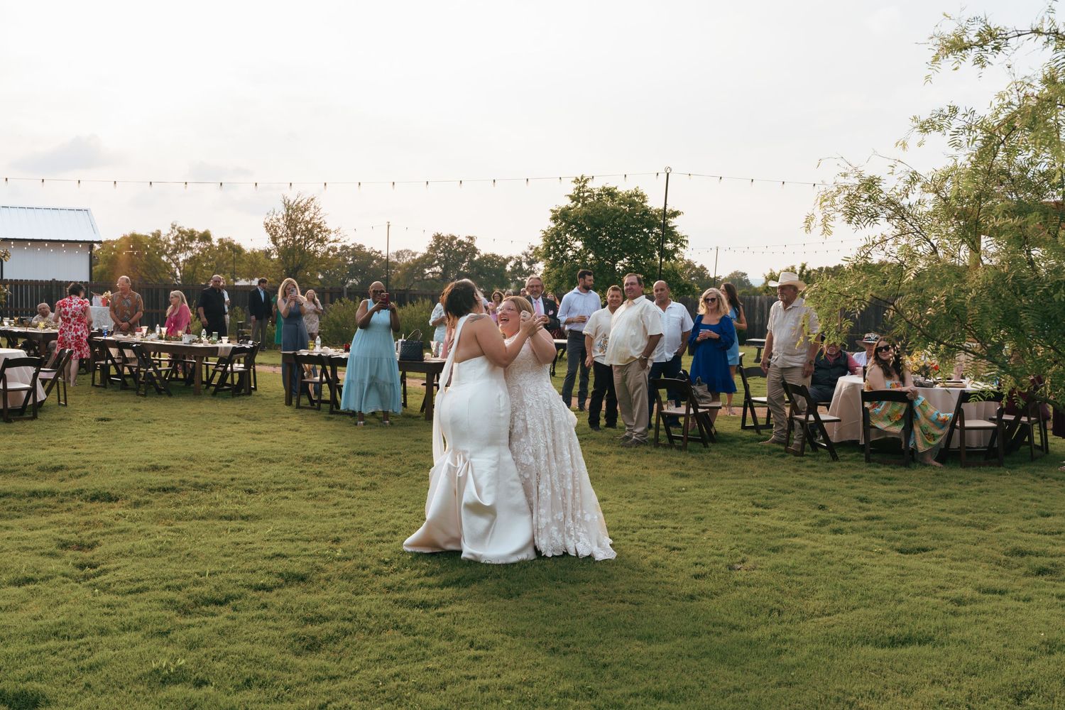Wedding guests gather on a grassy hillside during an outdoor reception at sunset.