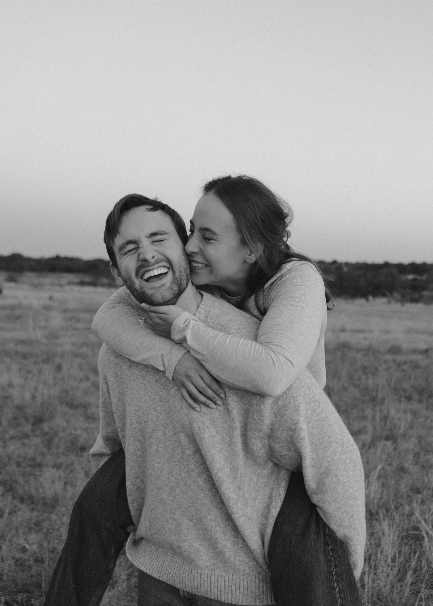 A joyful couple embraces in a field during a black and white photoshoot, with one person giving a piggyback ride.