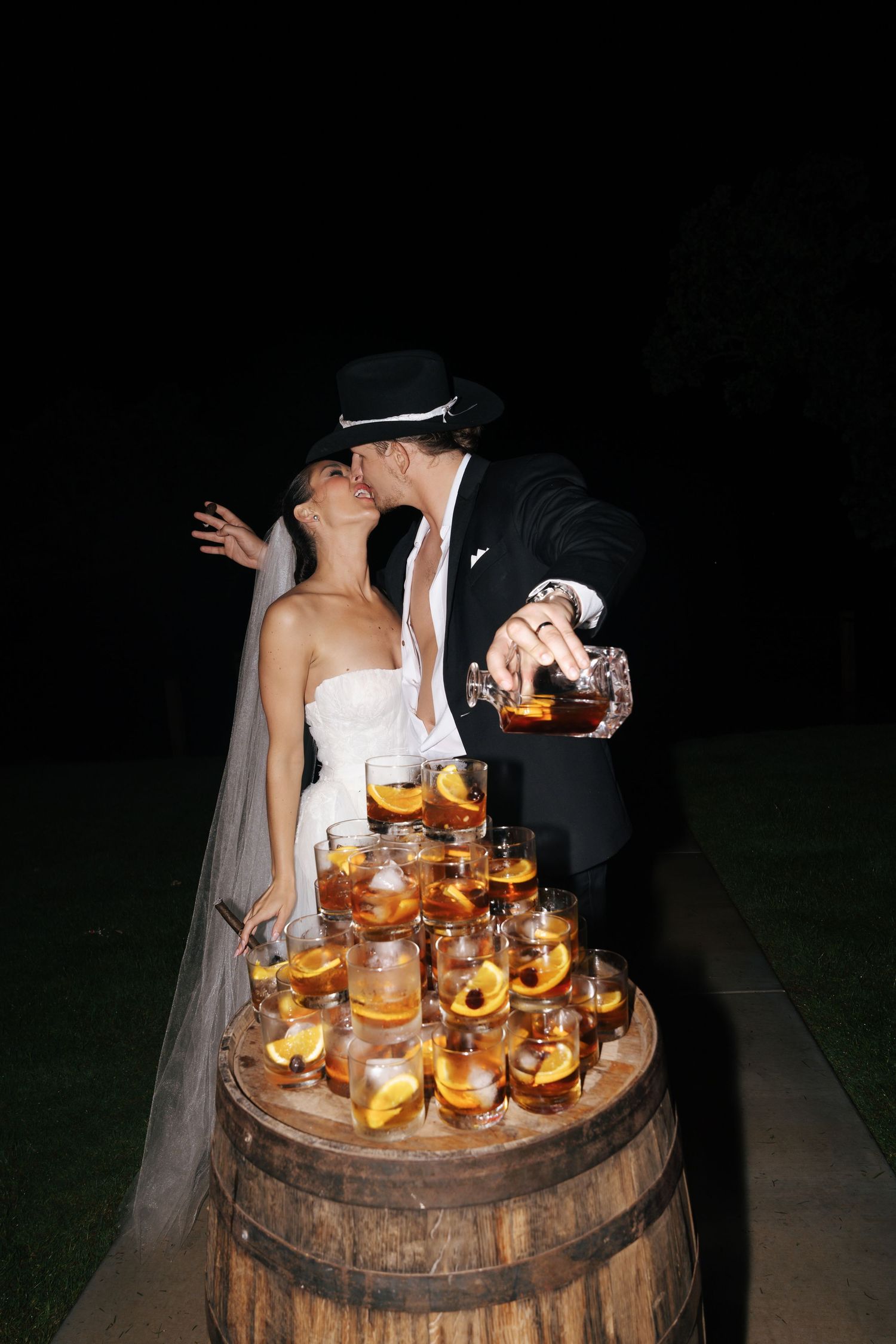 Couple celebrating with glowing cocktails displayed on a wooden barrel at night.