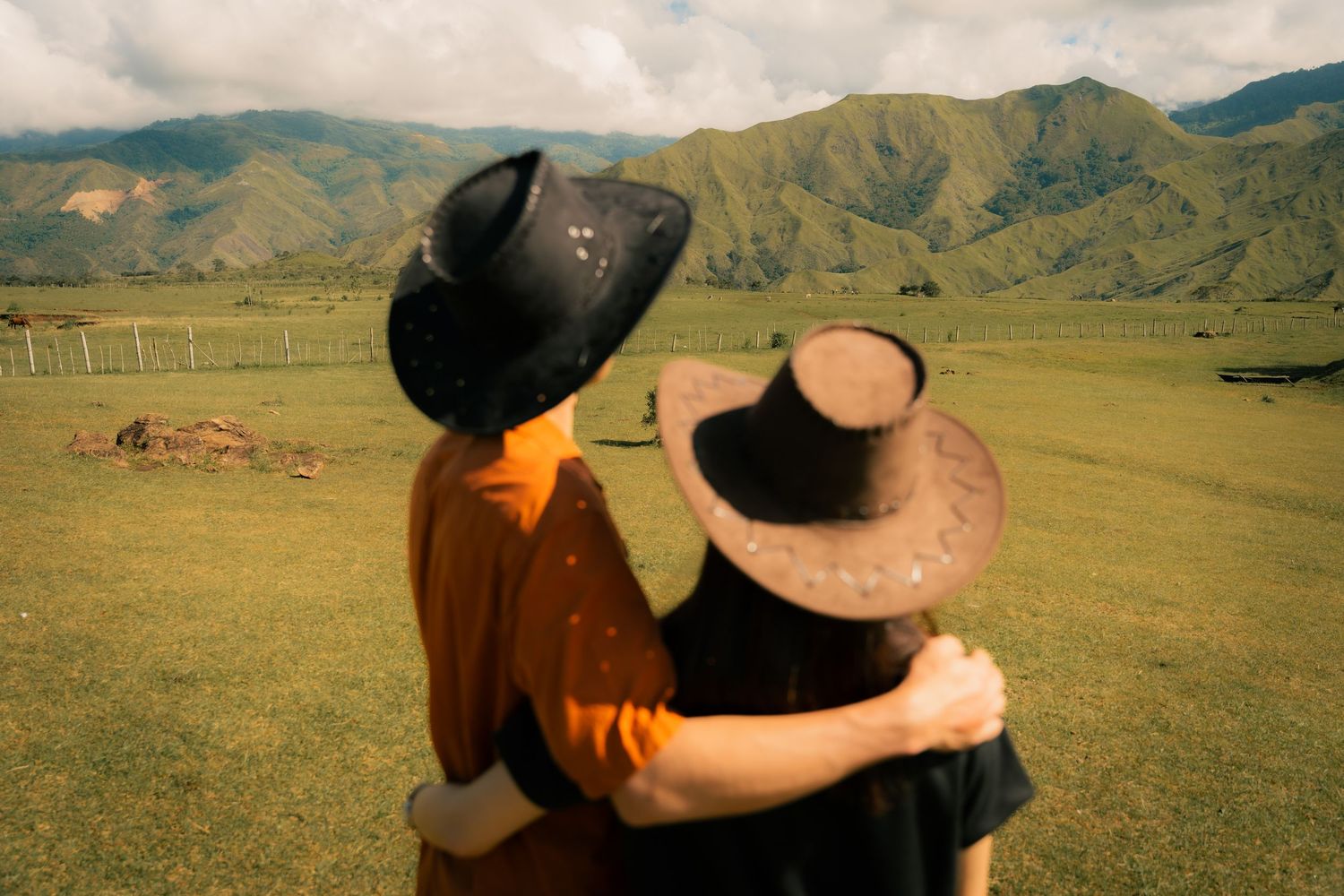 Two friends in cowboy hats embrace while overlooking a lush green valley with mountains in the background.