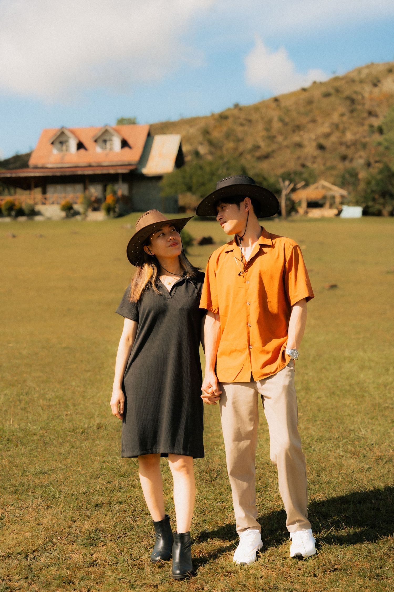 A couple in cowboy hats holds hands on a grassy field, with rustic buildings and a hill in the background.