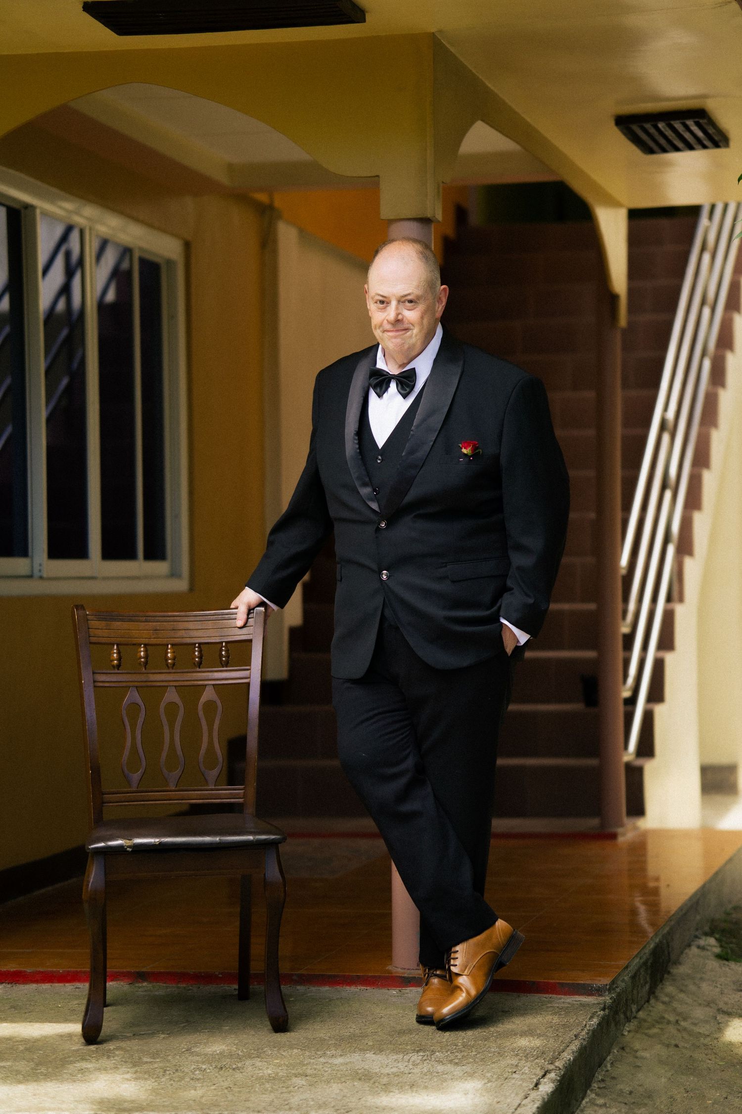 Elegant man in black tuxedo with bow tie and tan shoes posing beside a wooden chair on a porch.