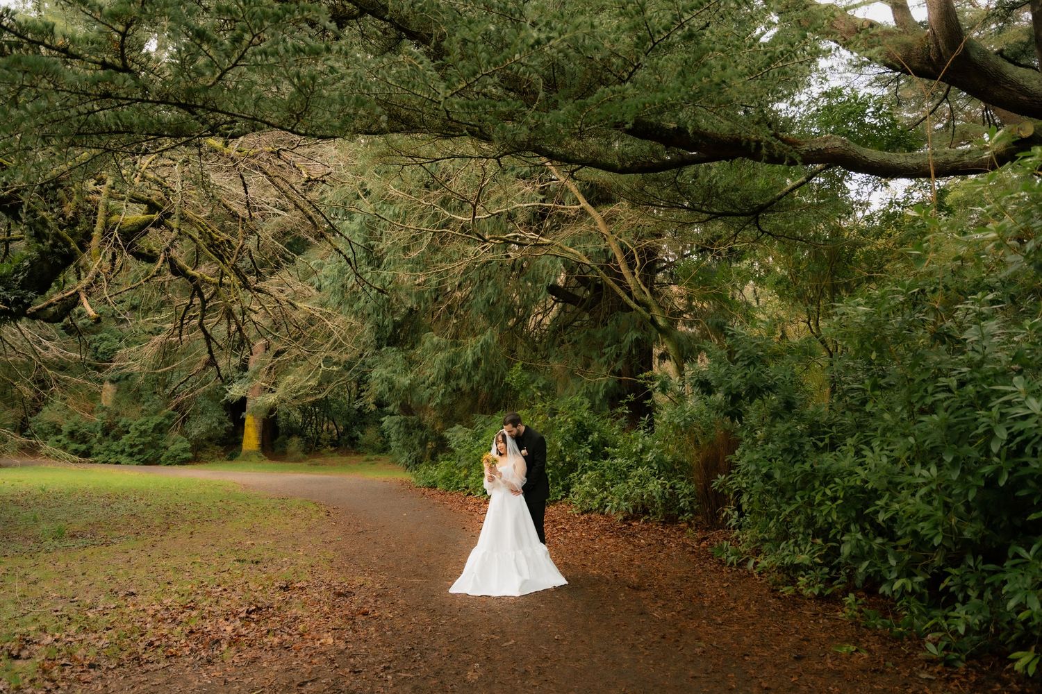 Bride and groom stand beneath sweeping moss-covered trees on a lush garden path in a romantic outdoor wedding portrait.