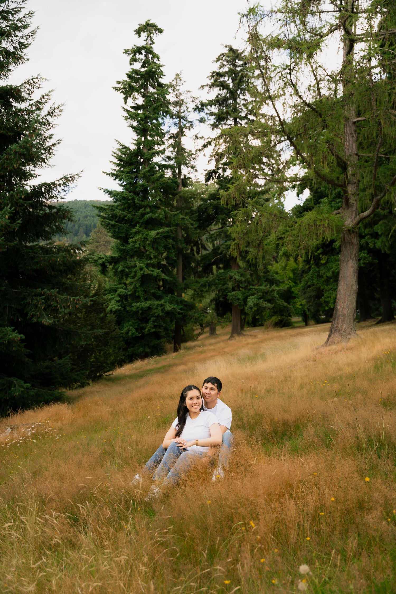 Couple sitting together on golden grass hillside surrounded by tall evergreen trees in a lush forest setting.