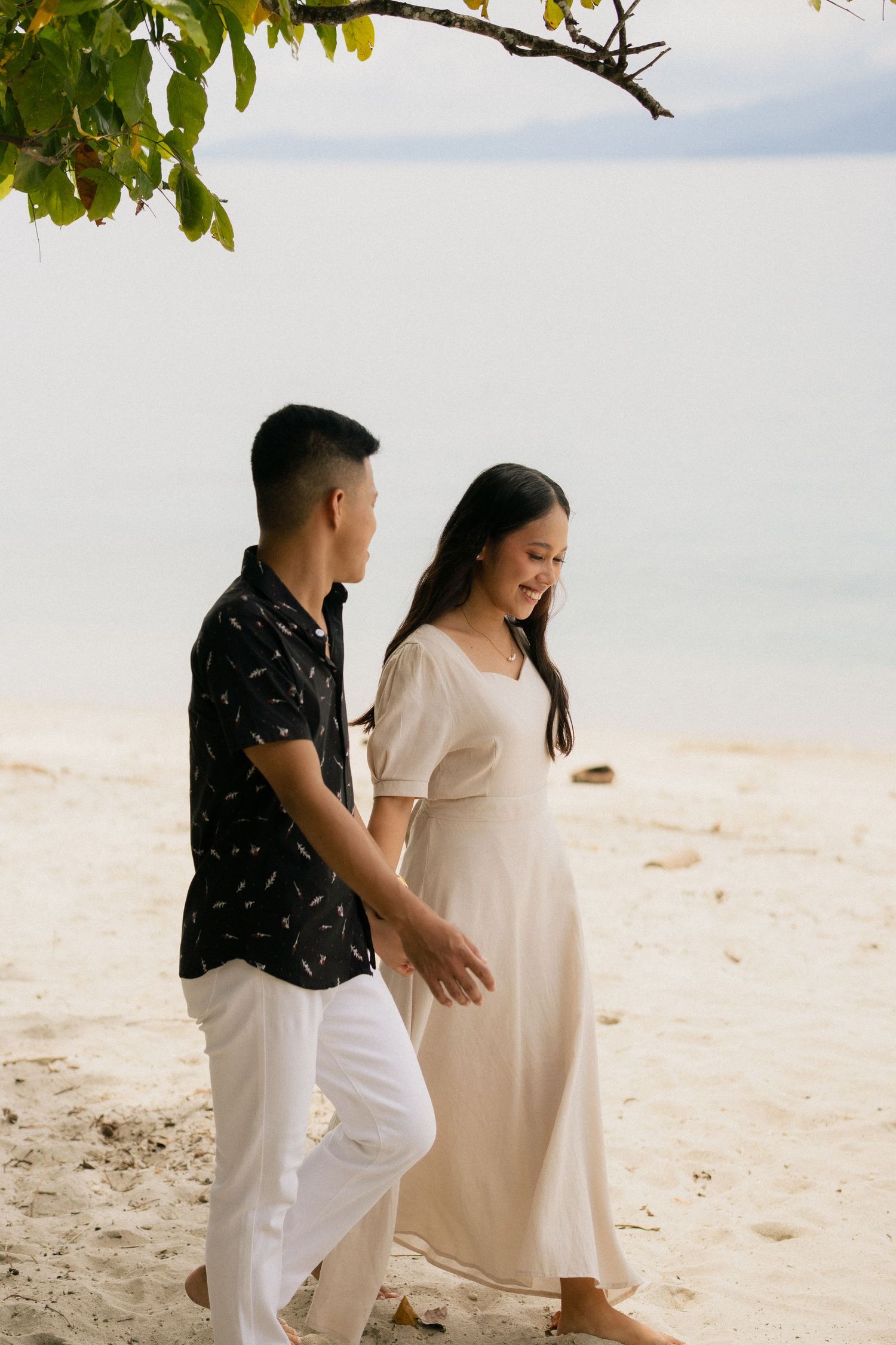 A couple walks hand in hand along a sandy tropical beach near calm waters beneath green tree branches.
