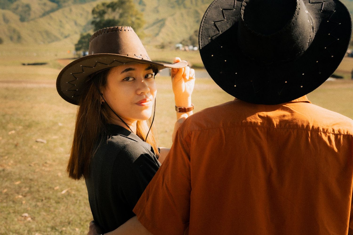 A young woman in a cowboy hat looks back over her shoulder outdoors, standing near a person in an orange shirt.