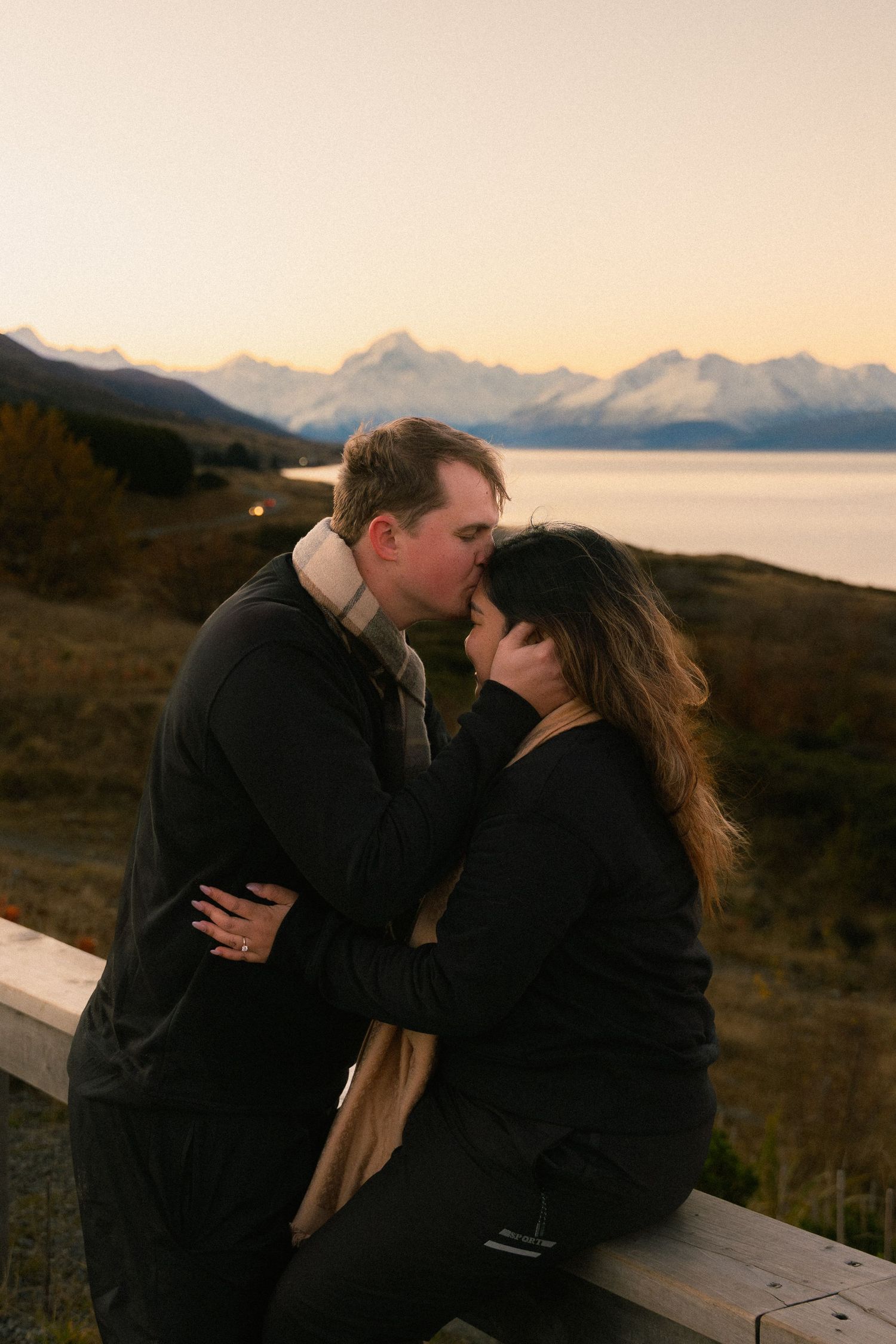 Couple sharing a romantic kiss at sunset with snow-capped mountains and lake in the background.