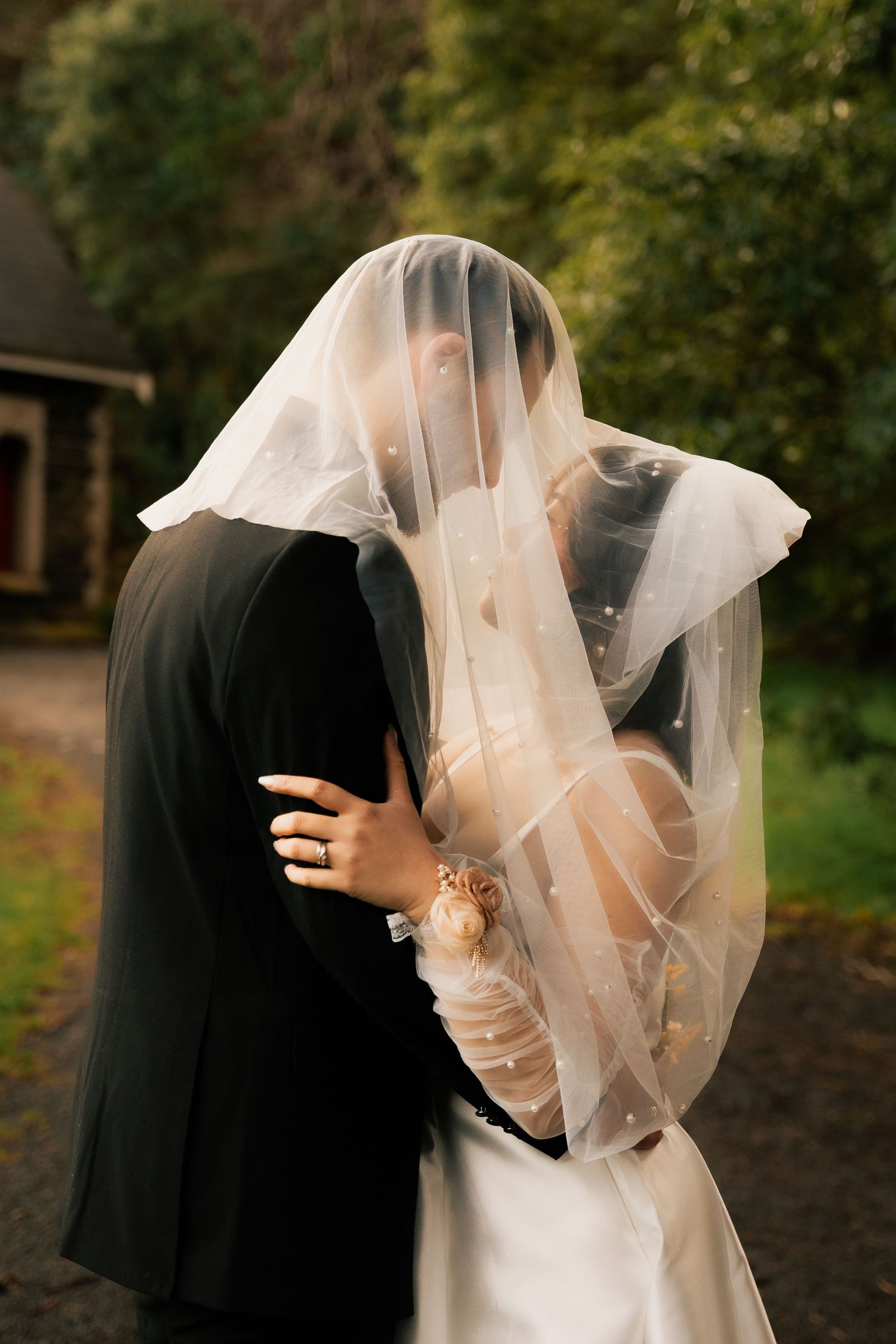 Bride and groom kissing under a sheer wedding veil outdoors, surrounded by lush green trees.