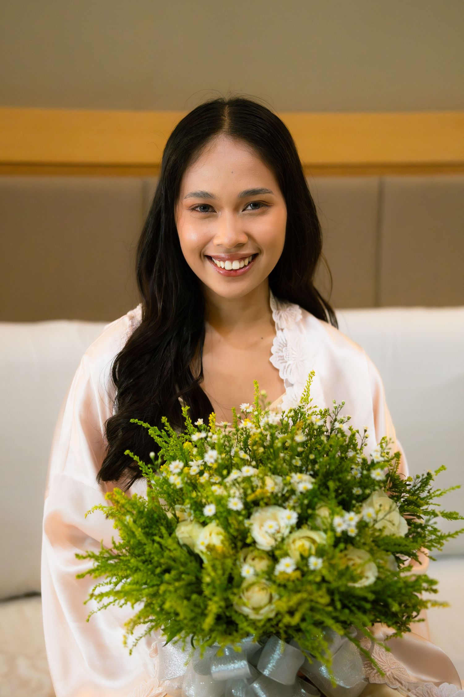 Smiling Asian bride in pink satin robe holding a lush bouquet of white roses and greenery in a hotel room.