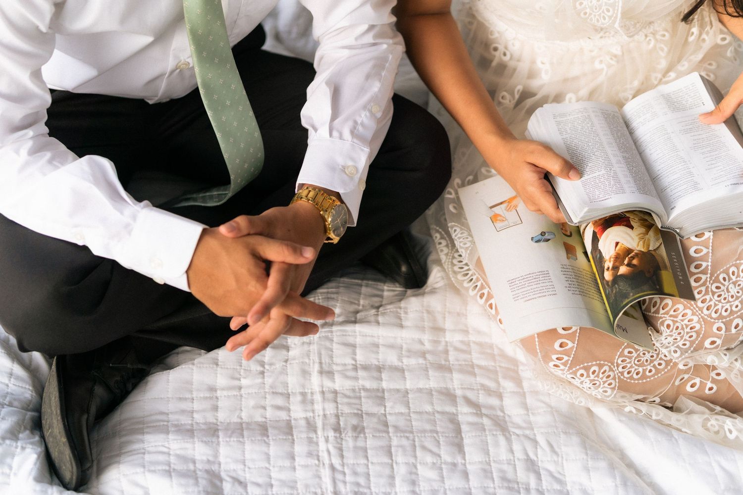 A couple in wedding attire sits on a white bed reviewing documents with a gold watch and sage tie visible.